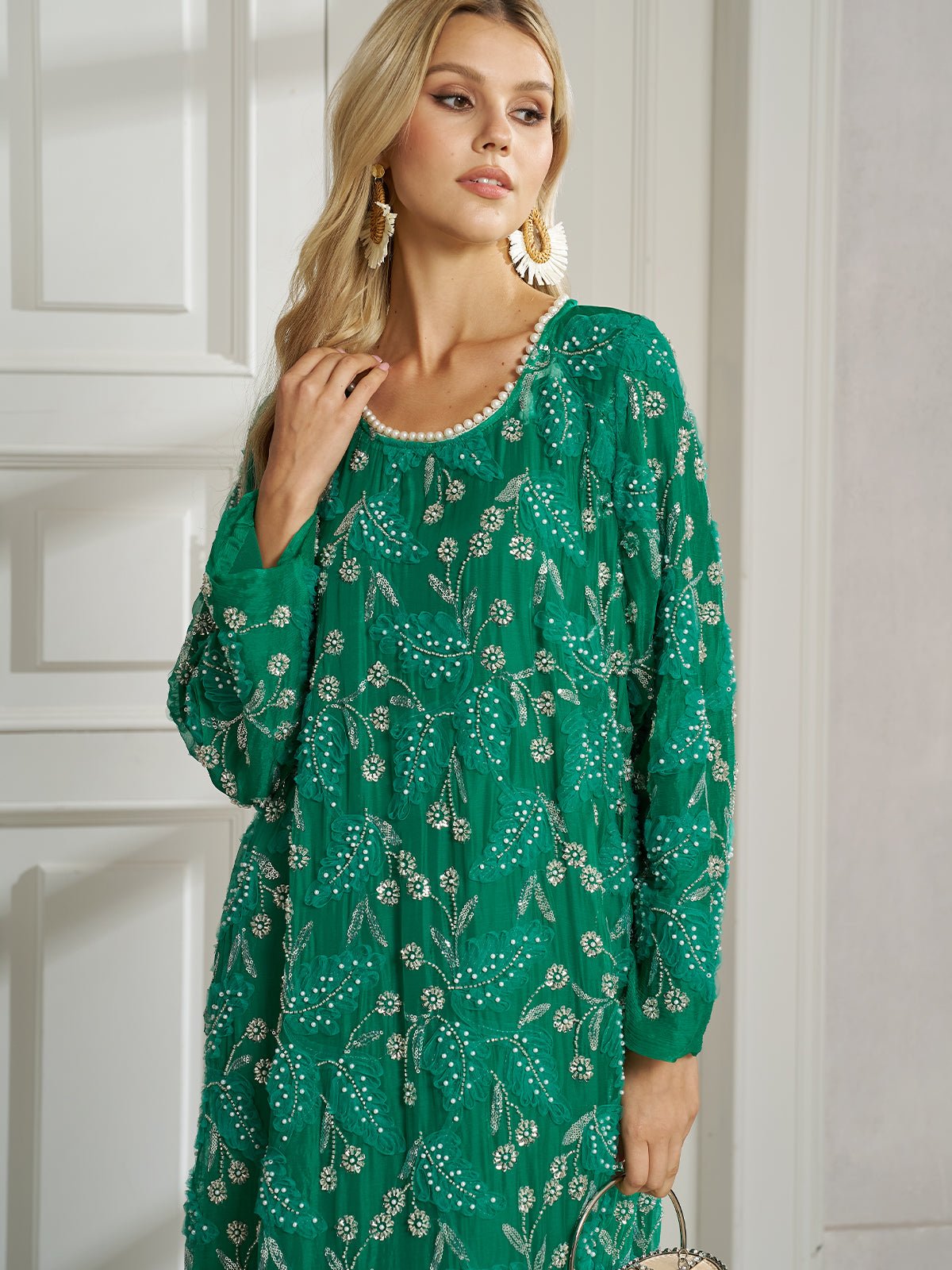 A woman indoors wears the Social Queen 100% Silk Beaded Embroidery Dress in green, featuring long sleeves and floral beading, paired with large gold earrings and a small handbag.