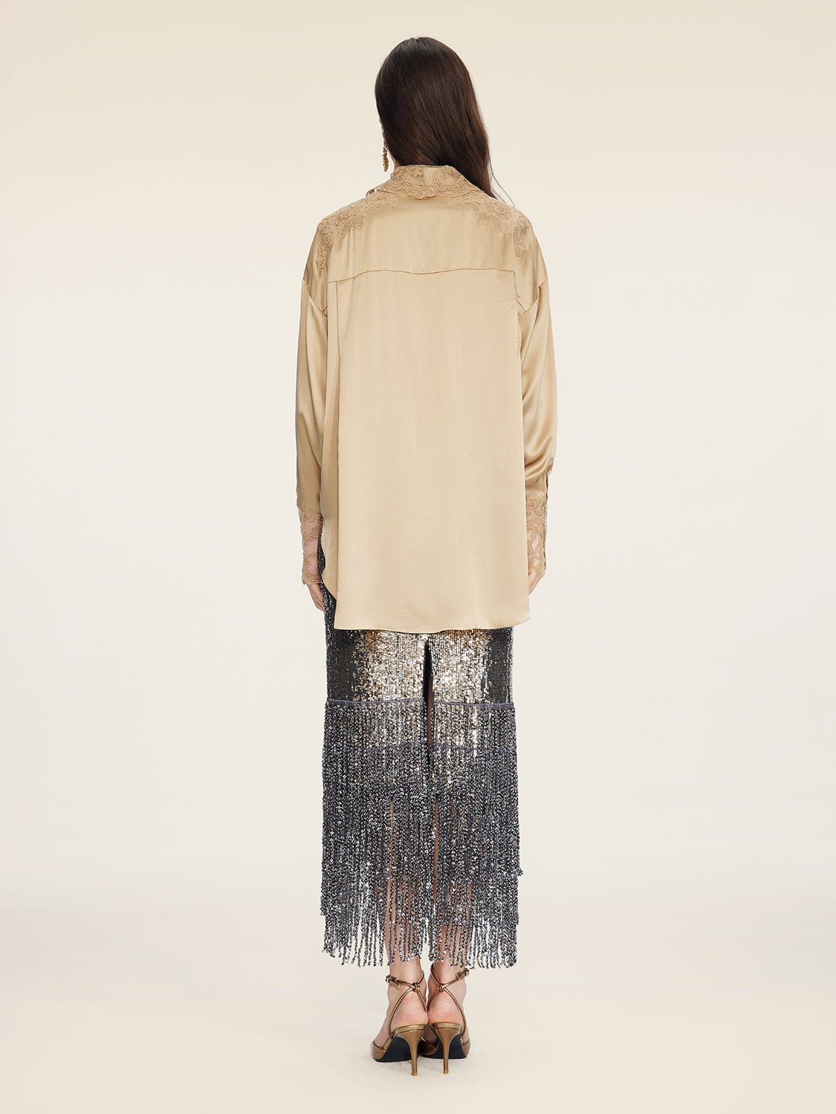 A person stands facing away, wearing the Social Queen 100% Silk Embroidered Lace Satin Blouse in beige, paired with a sequined silver fringe skirt and strappy heels against a plain light background.