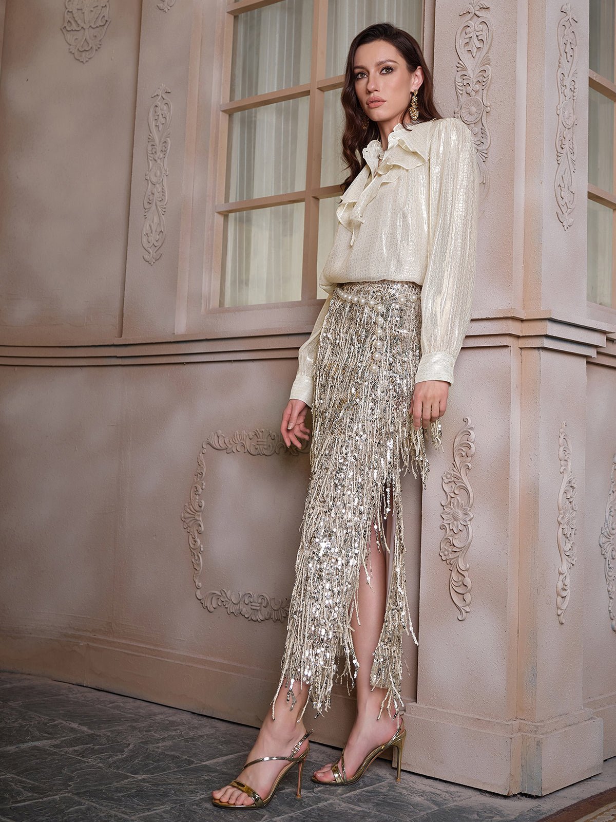 A woman stands indoors against an ornate wall, wearing the Social Queen 100% Silk Luminous Metallic Ruffle Blouse paired with a silver sequin fringed skirt and strappy heels.