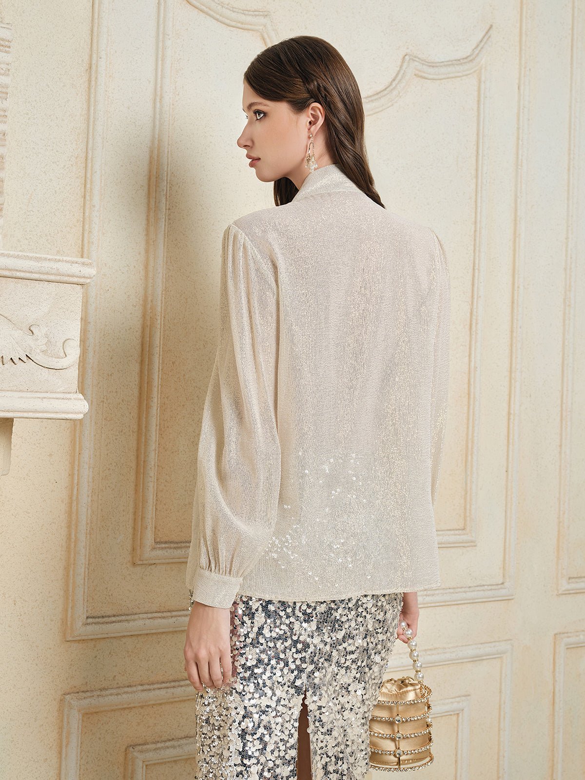 A woman stands indoors facing away, wearing the Social Queen 100% Silk Metallic Ribbon-Tie Blouse with puff sleeves and a sequined skirt. She holds a small, round handbag and wears drop earrings.