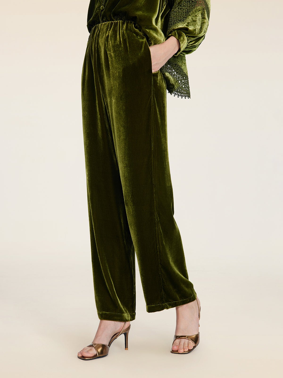 A person models Social Queen’s 100% Silk Velvet Wide-Leg Pants in vintage-inspired olive green, styled with metallic high-heeled sandals and a hand in their pocket, posed against a plain background.