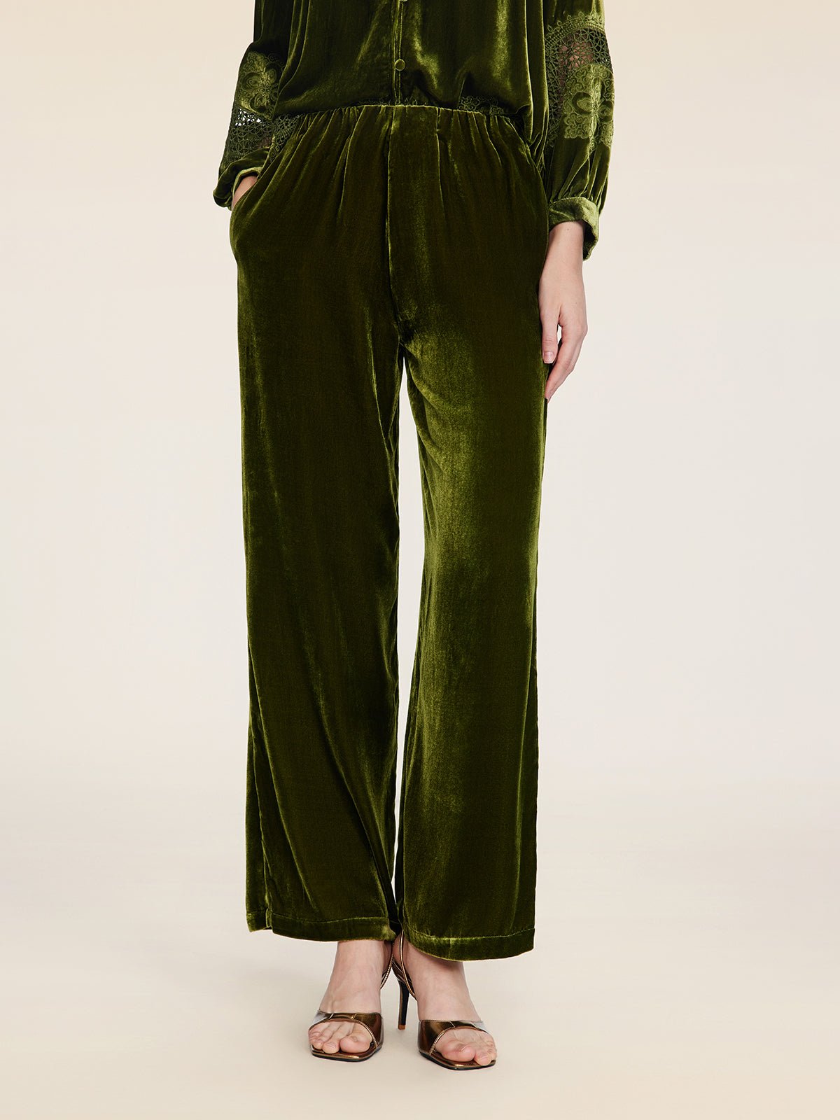 A person stands against a plain background wearing Social Queen’s 100% Silk Velvet Wide-Leg Pants in olive green with brown strappy heels, hands casually in their pockets.
