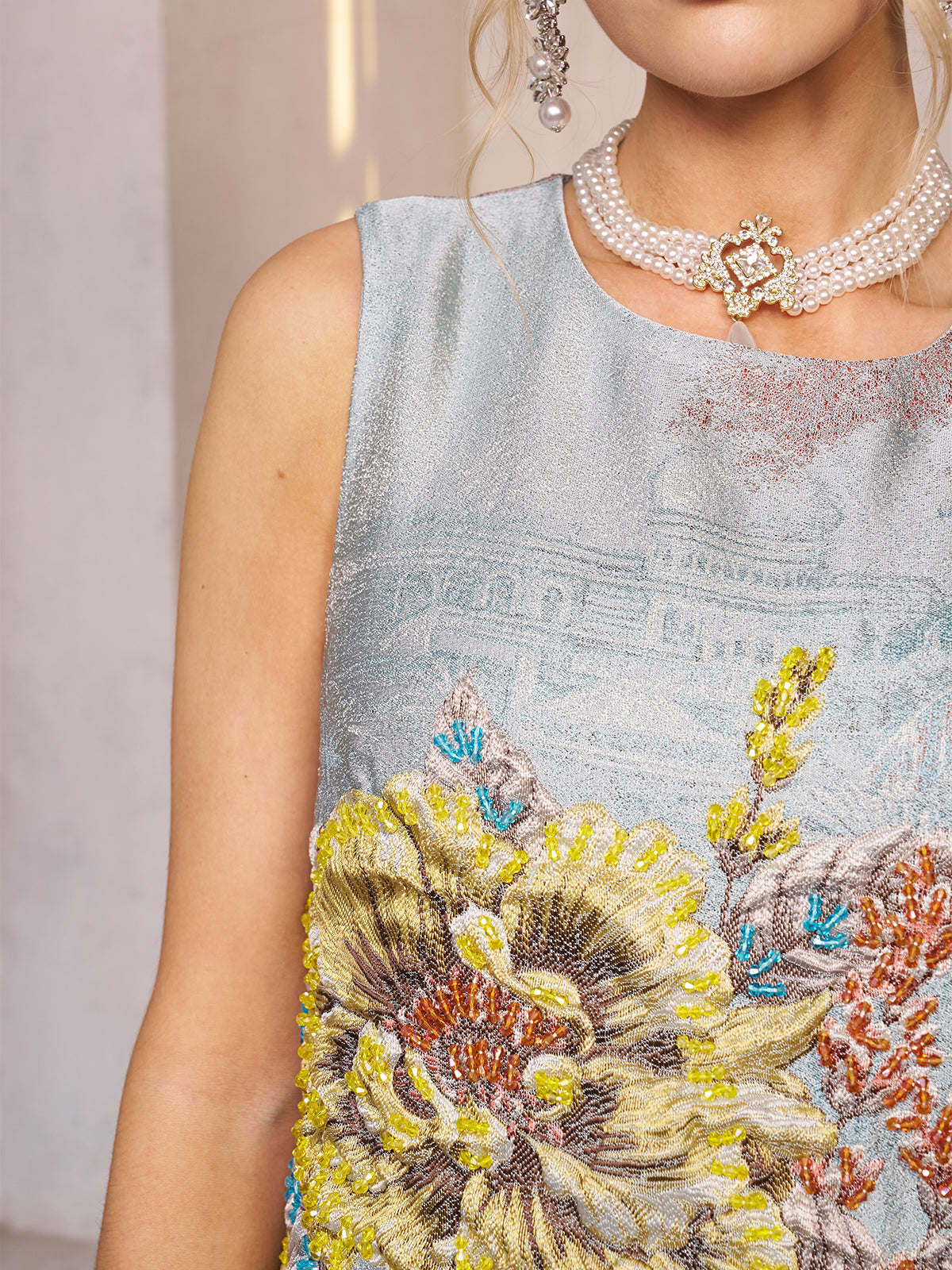 A woman in the Social Queen Floral Jacquard Beaded Sleeveless Dress, styled with a layered pearl heart pendant necklace and pearl earring—an elegant wedding guest look. Her face is partially out of frame.