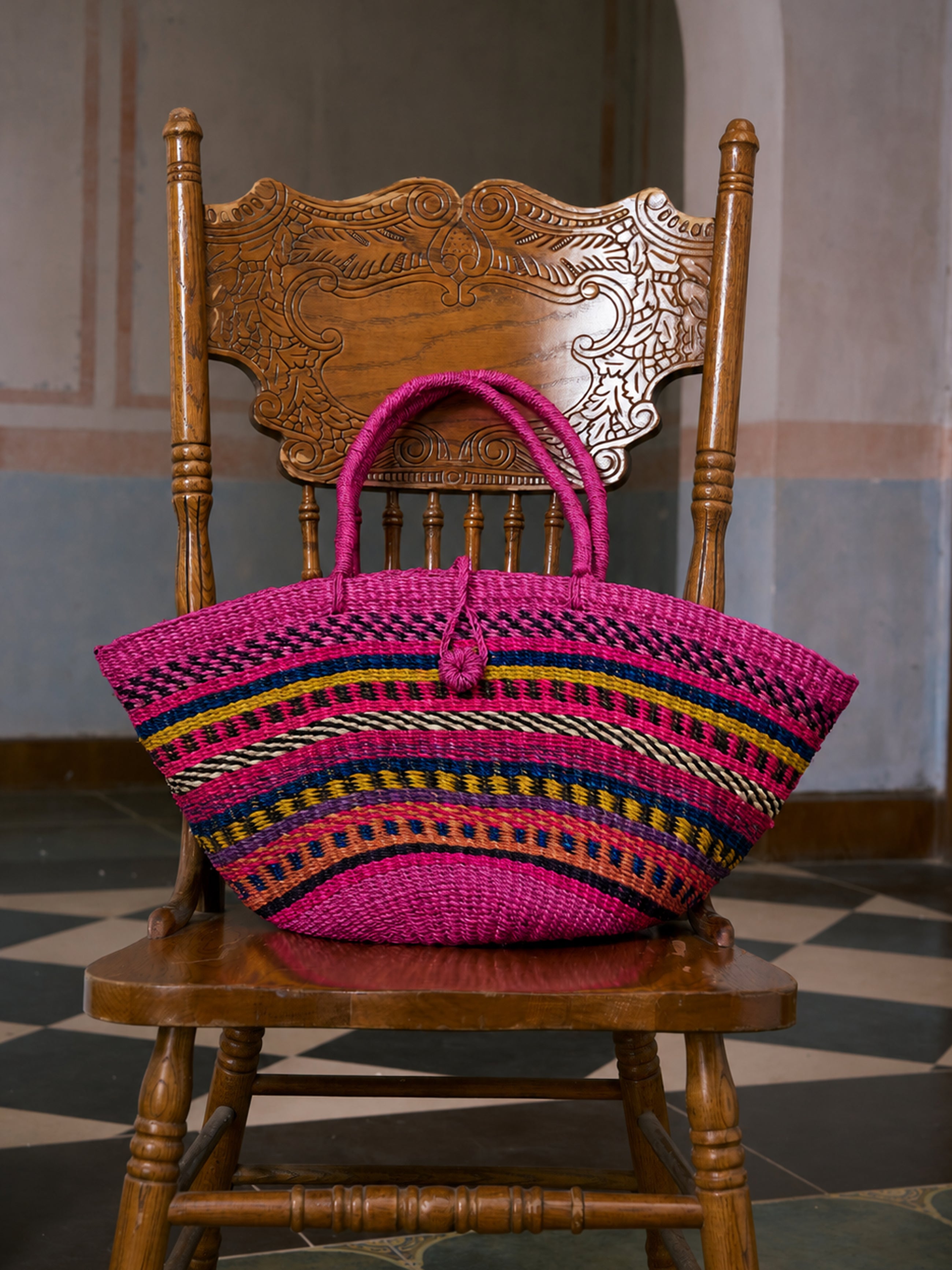 The Social Queen Colorful Striped Woven Tote Bag, handwoven in pink from natural ramie fiber and featuring multicolor striped accents, rests on an ornately carved wooden chair in a room with tiled floors and blue and pink walls.