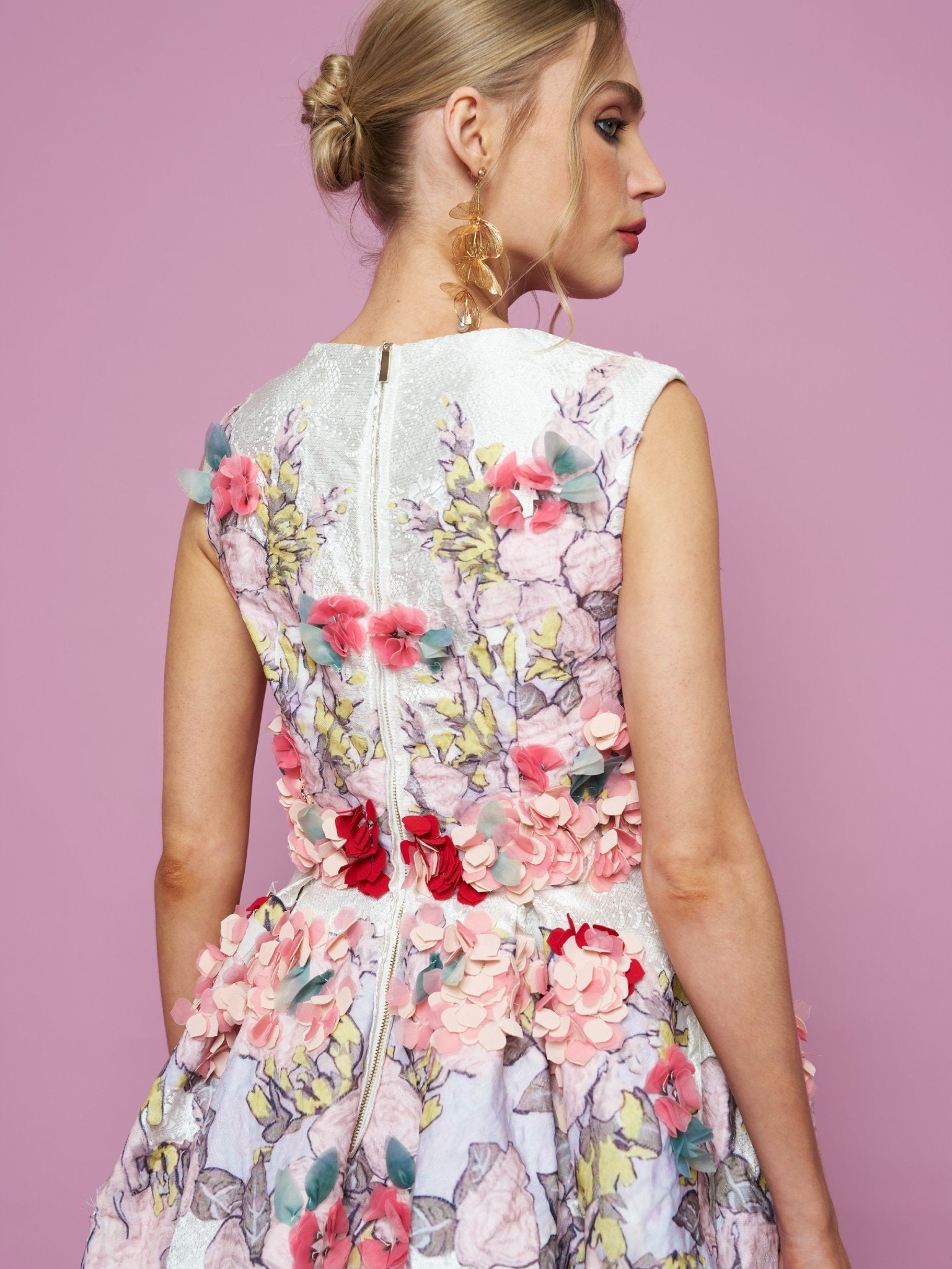 A woman faces away in the Social Queen 3D Floral Embellished Jacquard Gown, featuring sleeveless design, floral appliqué, and visible back zipper, posed against a pink background.