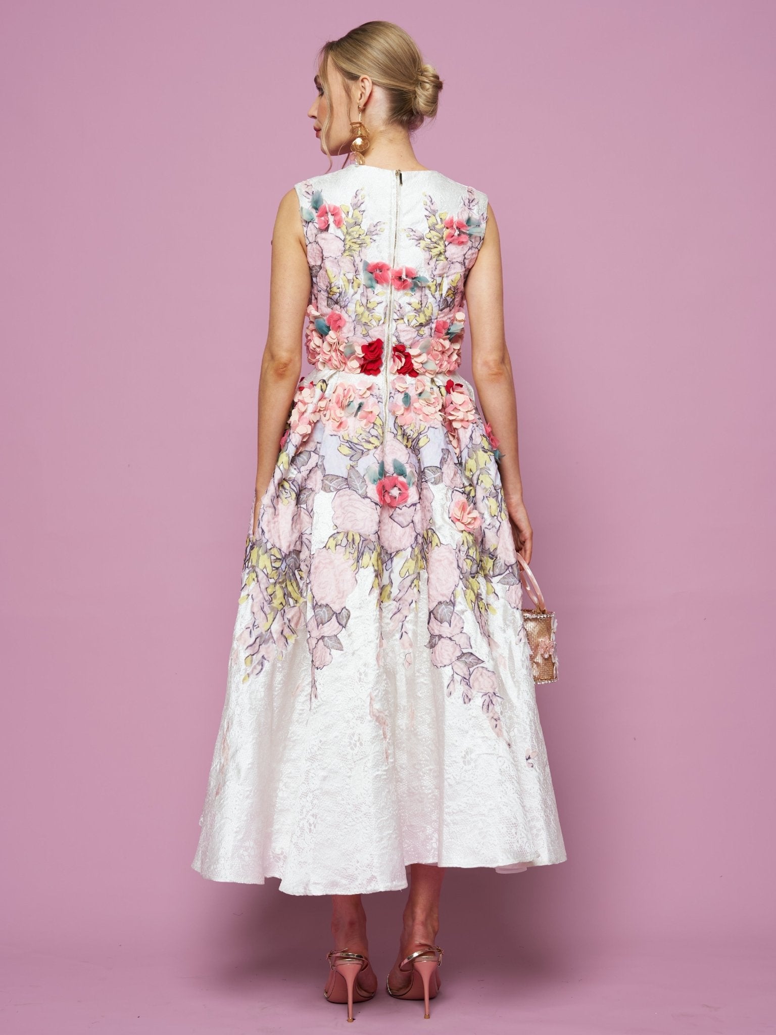 A woman stands against a pink background, facing away while holding a small handbag and wearing the Social Queen 3D Floral Embellished Jacquard Gown—sleeveless white satin with colorful 3D floral appliqué.