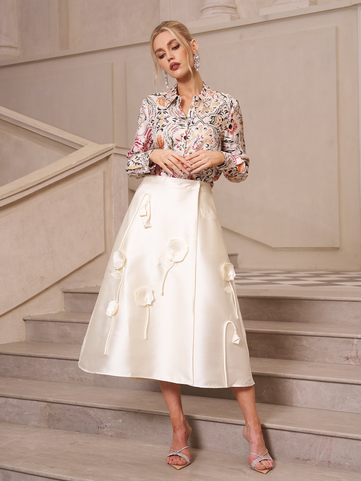 Woman stands on an indoor staircase wearing a floral blouse, strappy heels, and the Social Queen 3D Floral Satin Midi Skirt.