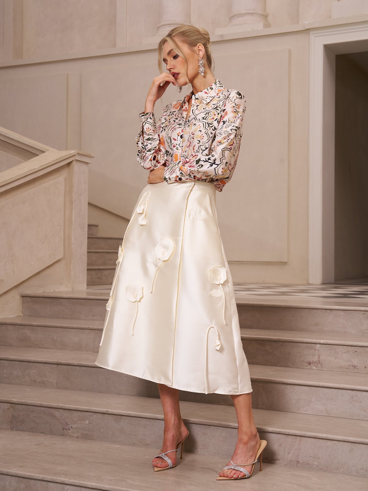 A woman stands on stairs in the Social Queen 3D Floral Satin Midi Skirt paired with a floral long-sleeve blouse and strappy sandals—a chic high waist A-line option for wedding guests.