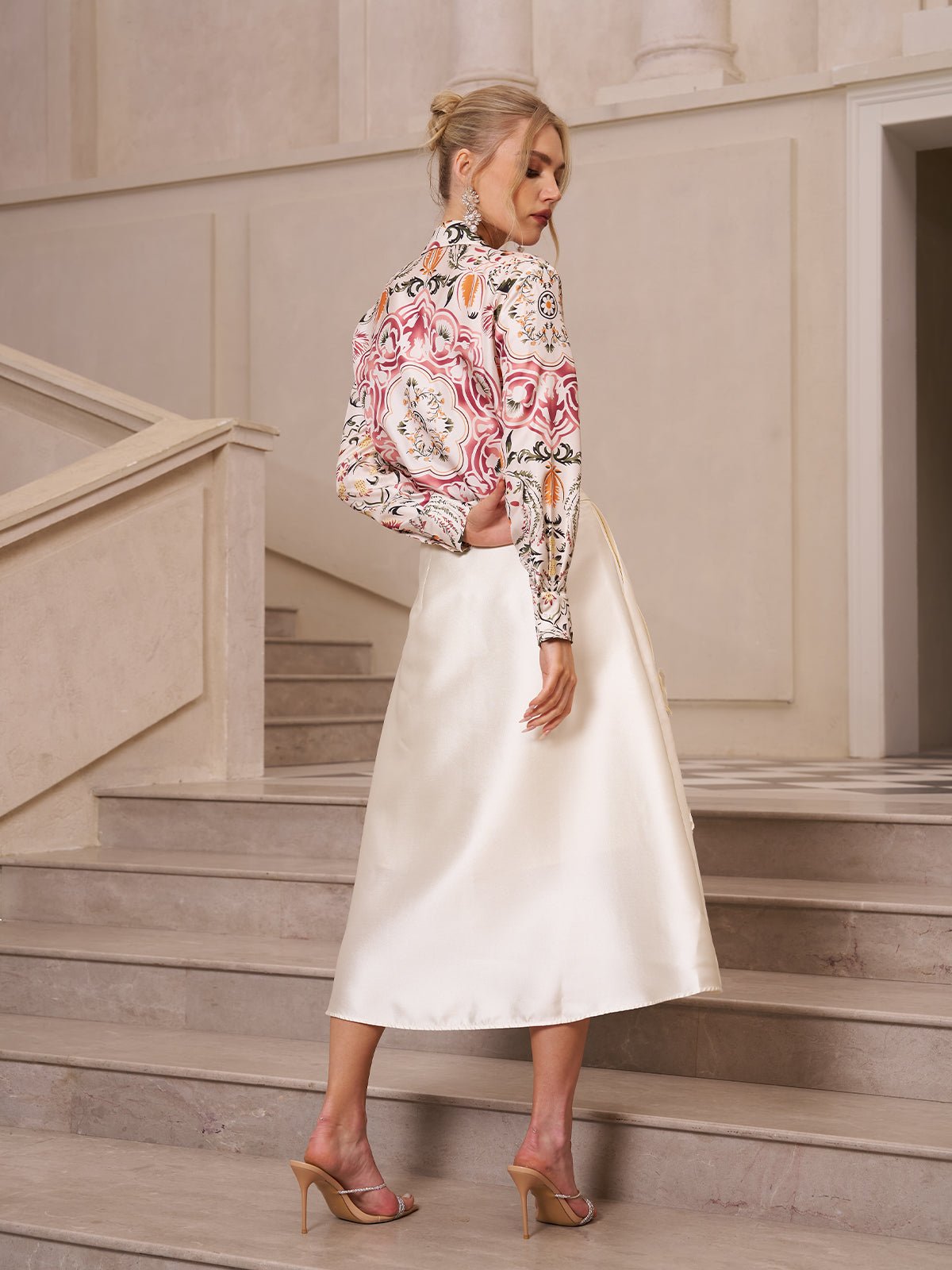 A woman stands sideways on stairs indoors, wearing a patterned long-sleeve top, heeled sandals, and the Social Queen 3D Floral Satin Midi Skirt.