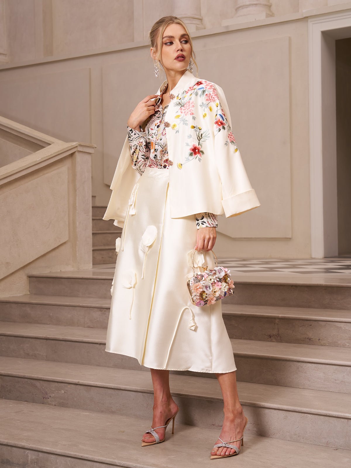 A woman stands on a staircase wearing the Social Queen 3D Floral Satin Midi Skirt with a cream floral embroidered coat, holding a small floral handbag and wearing heeled sandals.