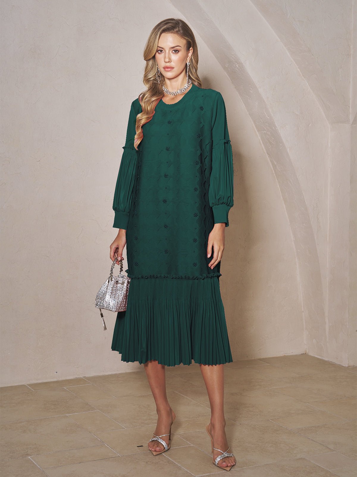 A woman stands indoors wearing the Beaded Jacquard Pleated Midi Dress by Social Queen, styled with silver sandals, a pearl-detailed statement necklace, and a silver handbag.