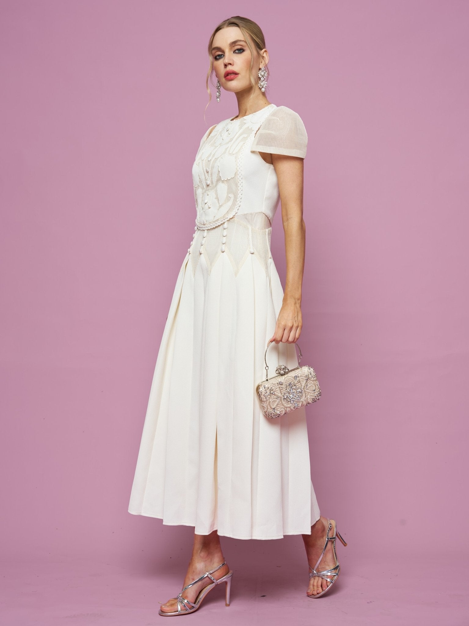 A woman wears the Social Queen Appliqué Pearl Waffle Mesh Dress with silver high-heeled sandals and an ornate small handbag, standing against a plain pink background.