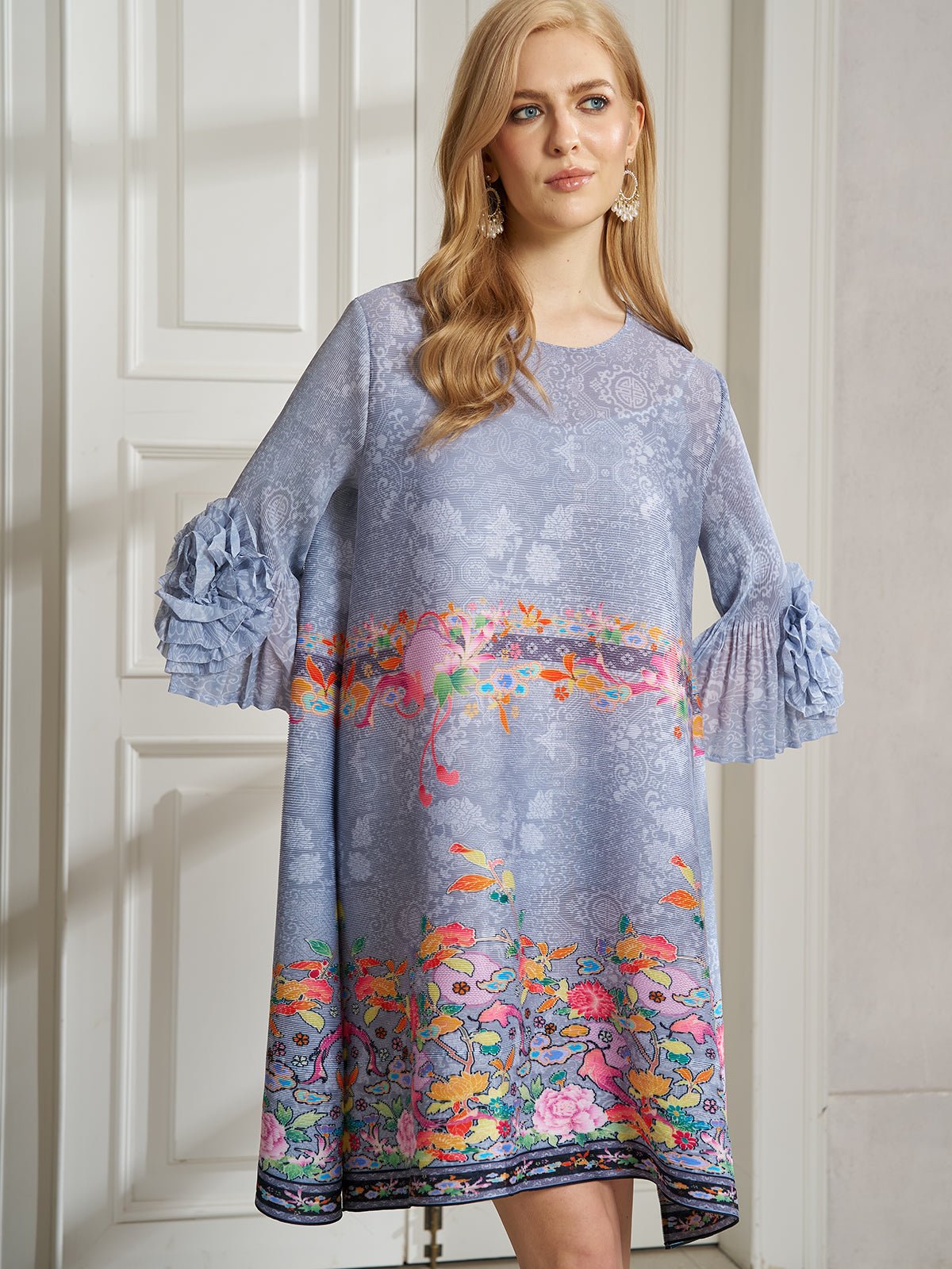 A woman stands indoors in front of white panel doors wearing the Social Queen Art-Print Floral-Appliqué Pleated Dress, featuring light blue floral patterns and ruffled sleeves.