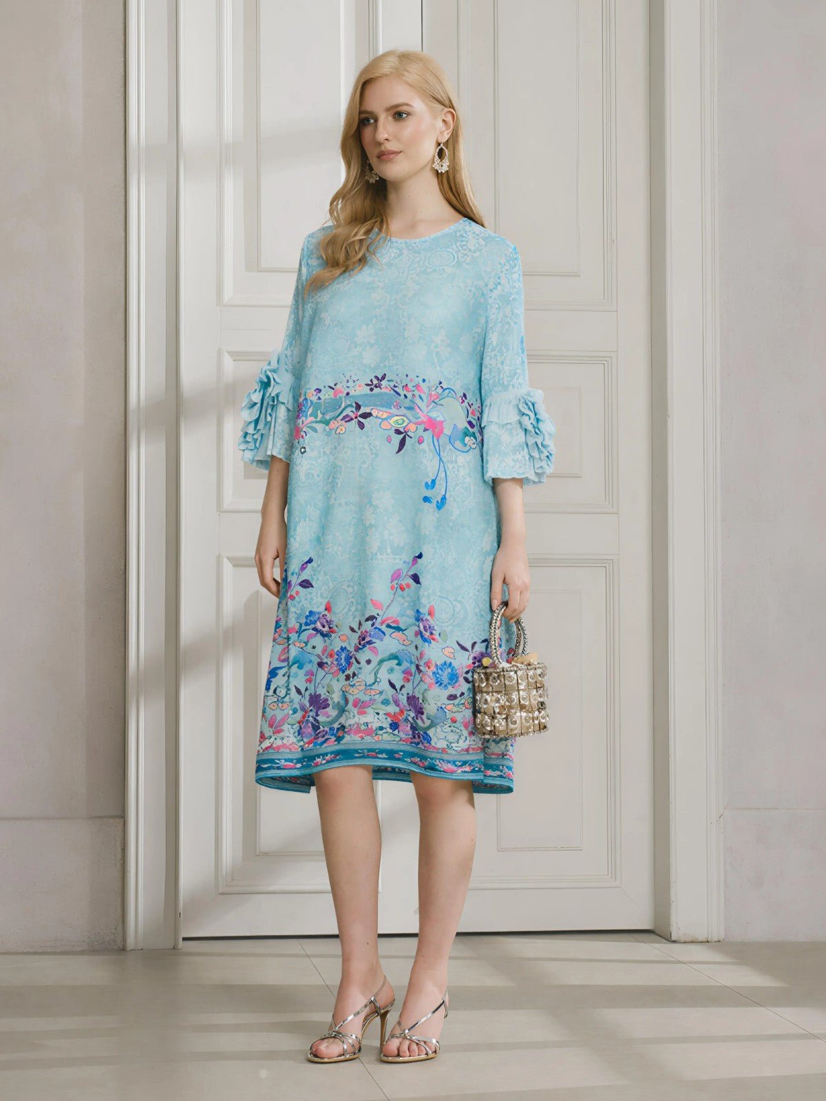 A woman stands indoors wearing the Social Queen Art-Print Floral-Appliqué Pleated Dress with ruffled sleeves, silver sandals, and a small handbag.
