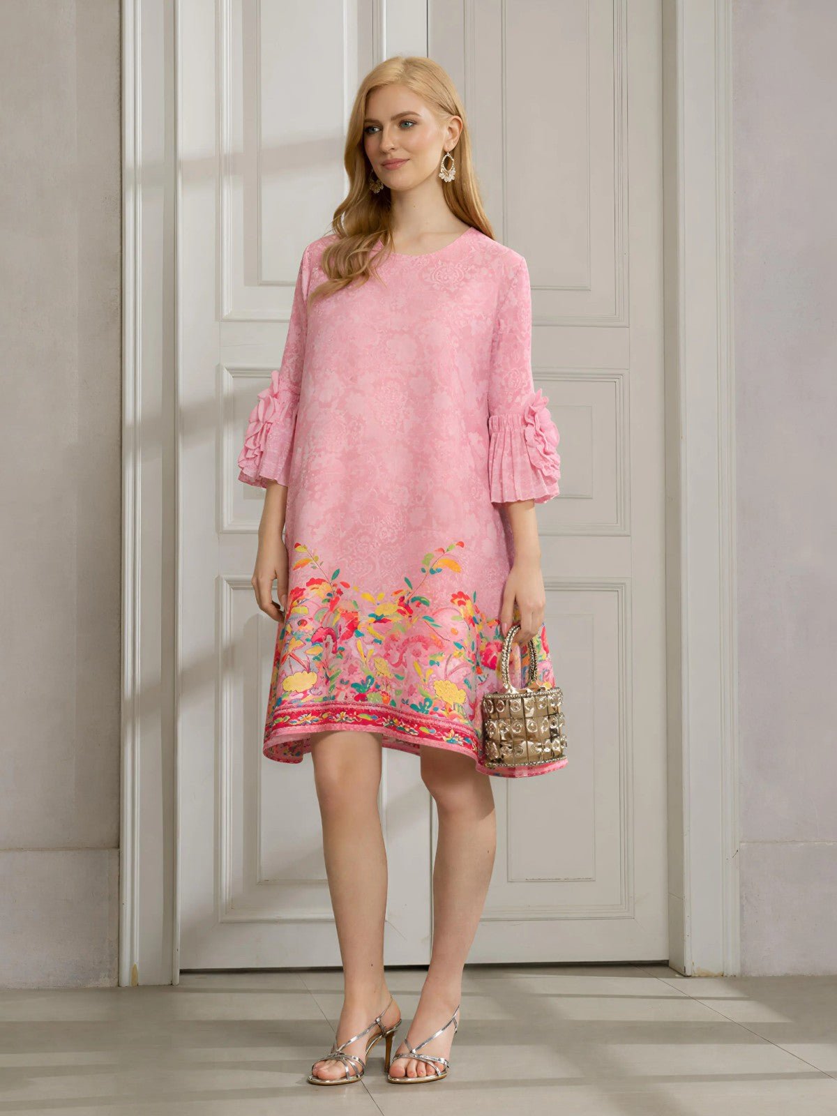 A woman stands indoors wearing the Social Queen Art-Print Floral-Appliqué Pleated Dress in light pink with ruffled sleeves, paired with silver sandals and a small metallic handbag.
