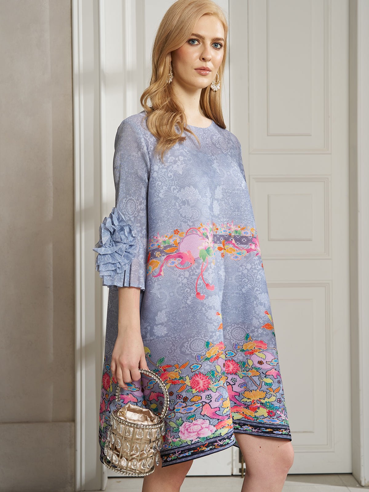 A woman stands indoors holding a small metallic handbag while wearing the Social Queen Art-Print Floral-Appliqué Pleated Dress with ruffled sleeves.