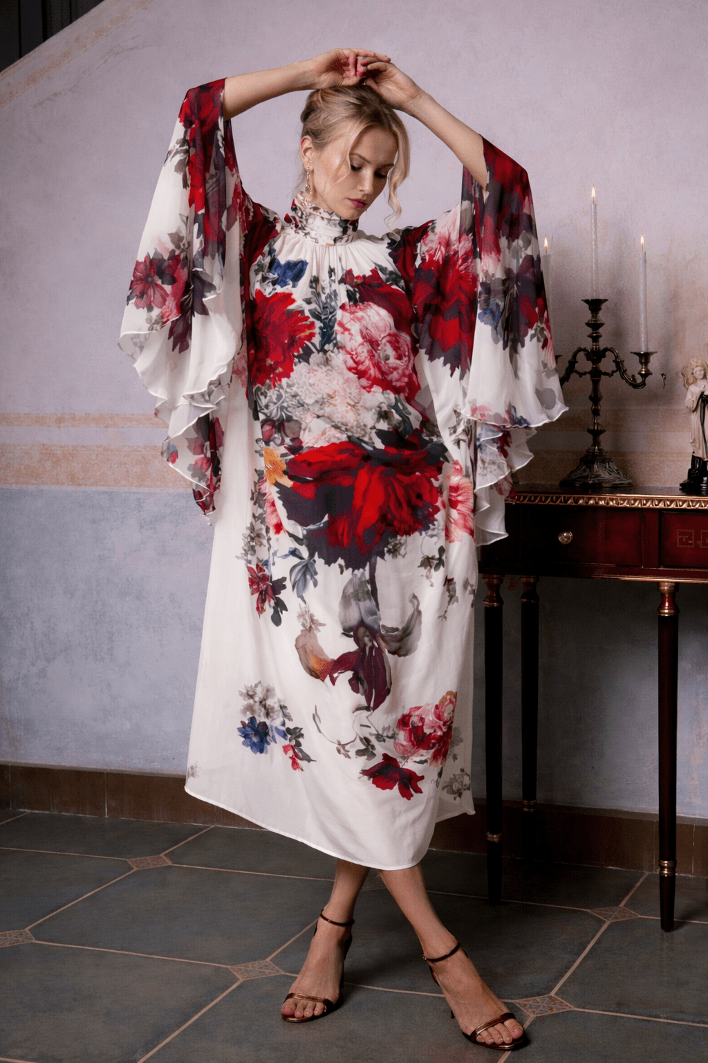 A woman wears the Social Queen Artistic Floral Print Ruffle Dress with wide, flowing sleeves indoors—arms raised near a dark wooden table and candelabra—making it a chic choice for wedding guests.