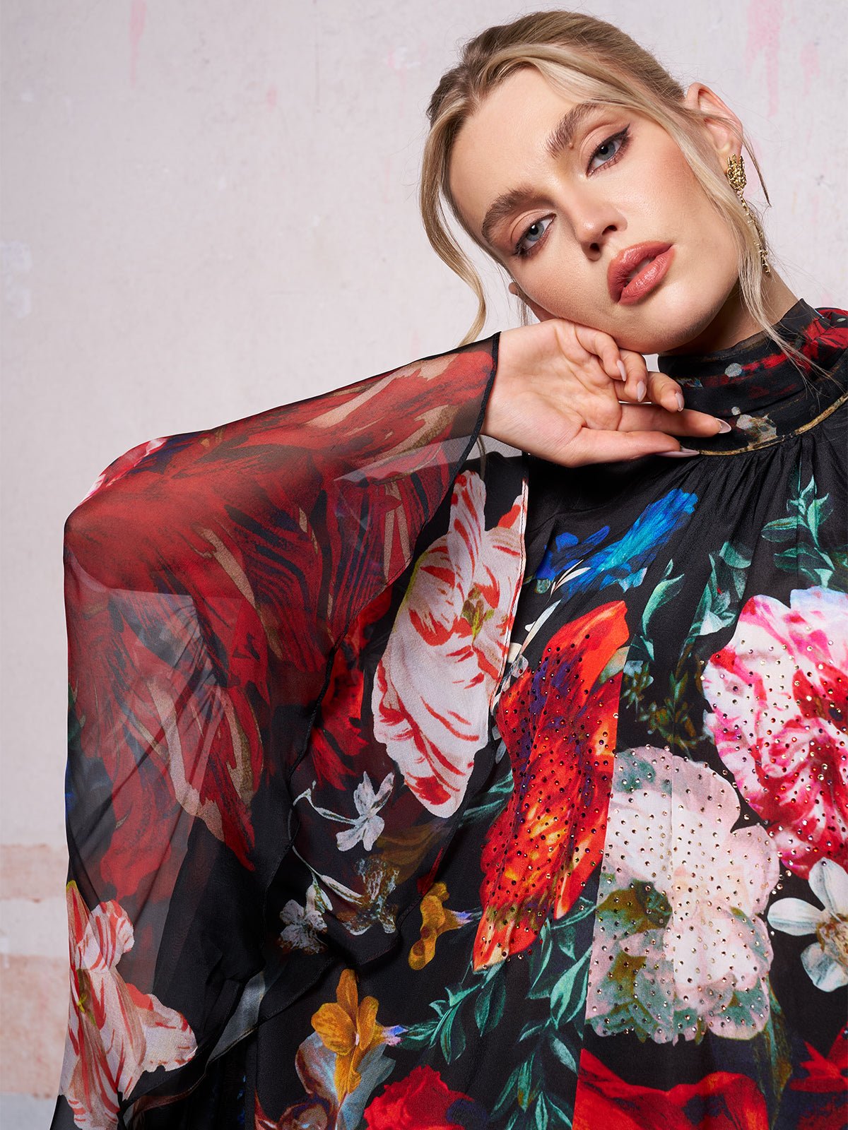 A woman models the Social Queen Artistic Floral Print Ruffle Dress, featuring sheer fabric with bold, colorful patterns, against a light-colored wall.
