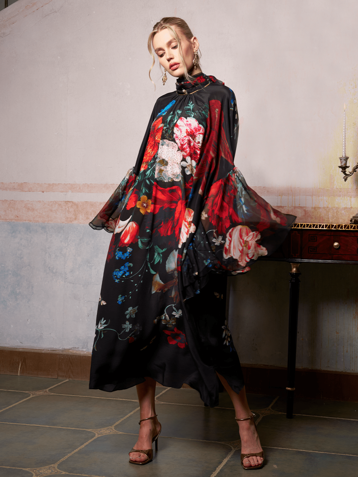 A woman poses indoors in the Social Queen Artistic Floral Print Ruffle Dress, featuring a long, flowing silhouette and vibrant floral patterns, standing beside a table with a single candleholder.