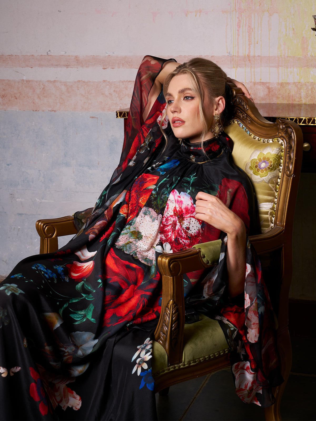 A woman in the Social Queen Artistic Floral Print Ruffle Dress sits on an ornate chair, her arm draped over the back, gazing to the side in a vintage-inspired room.