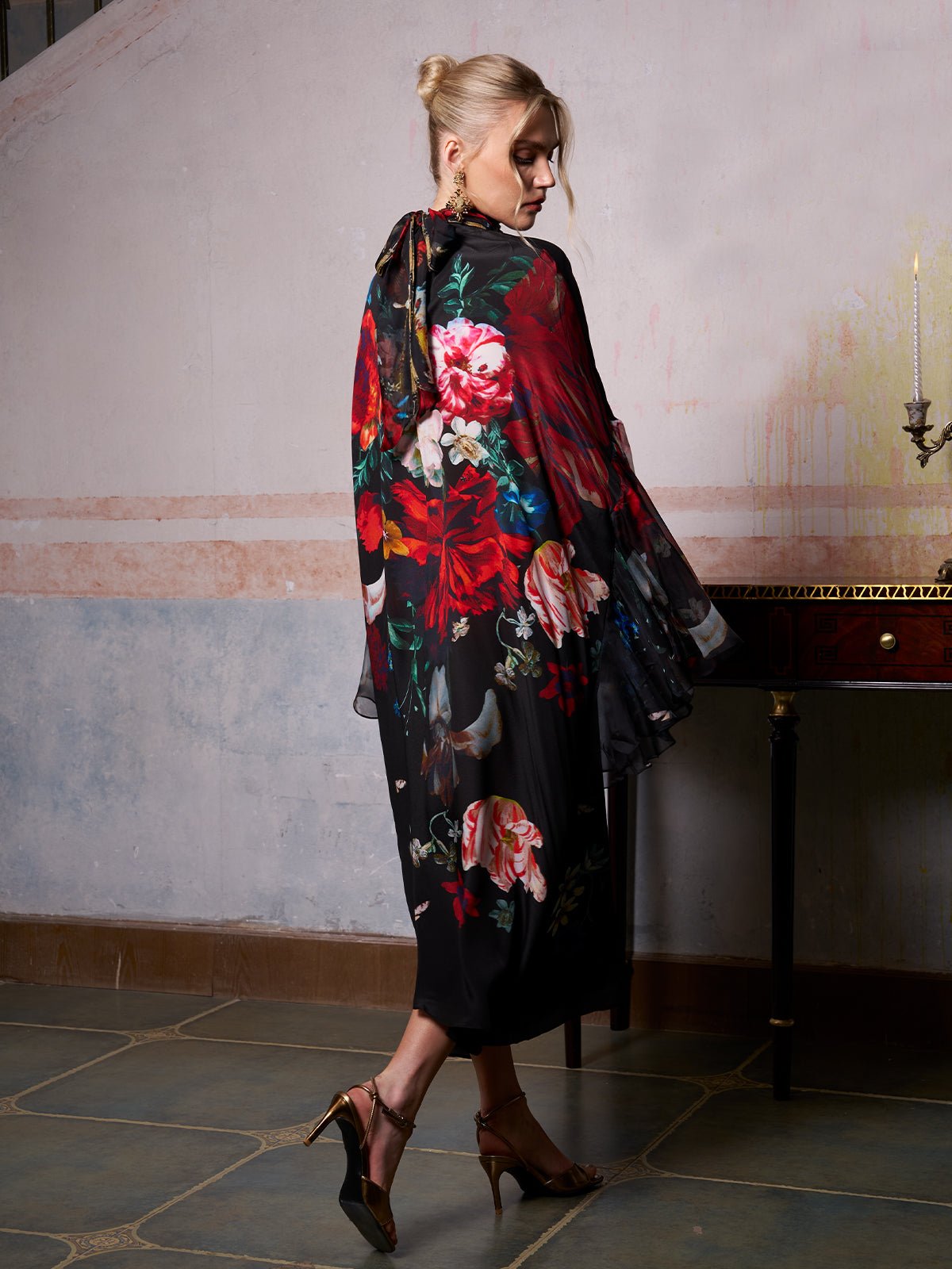 A woman in the Social Queen Artistic Floral Print Ruffle Dress and gold heels stands indoors, facing away from the camera near a wooden table with a brass candlestick.