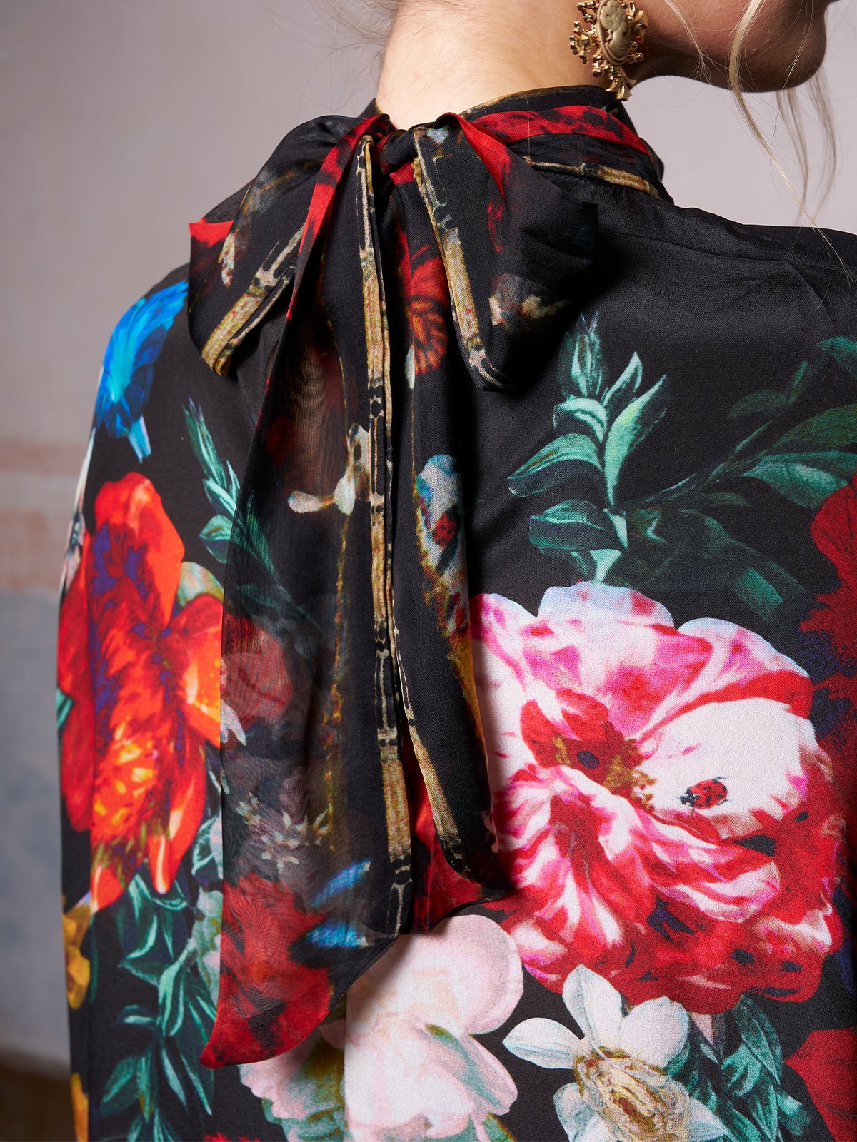 The image features the back of a person in the Social Queen Artistic Floral Print Ruffle Dress, a chic black dress with red, pink, and white blooms and a statement neck bow—ideal for a stylish wedding guest look.