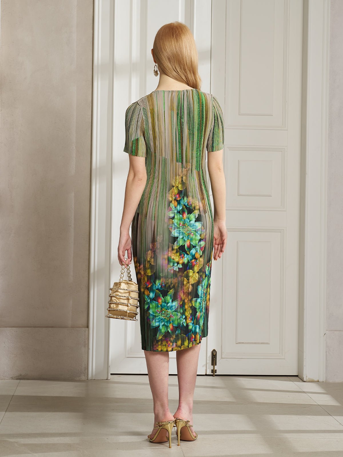 A woman with long red hair stands by a white door, wearing Social Queen's Artistic Smudge Print Plissé Dress and holding a small gold handbag—an elegant choice for chic resort style.