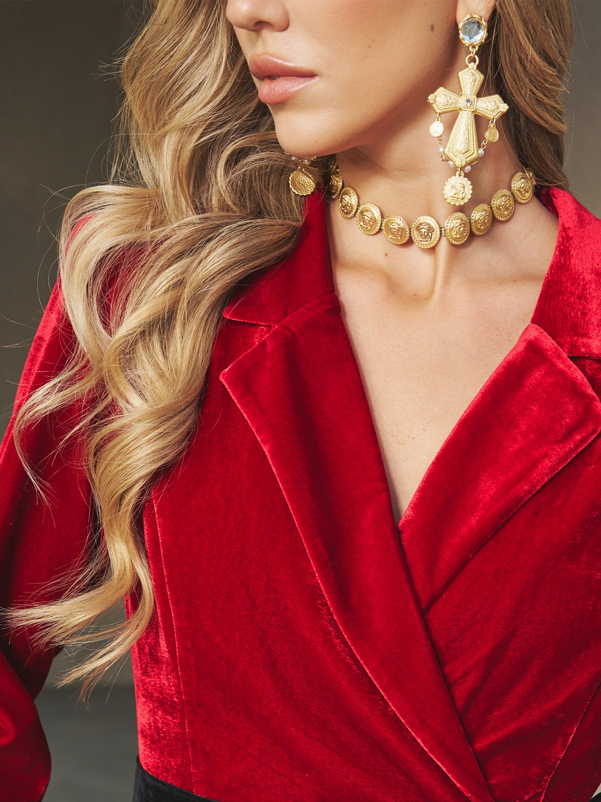 A woman with long wavy blonde hair wears a red velvet blazer, the Social Queen Baroque Medallion Necklace, and a large gold cross earring.