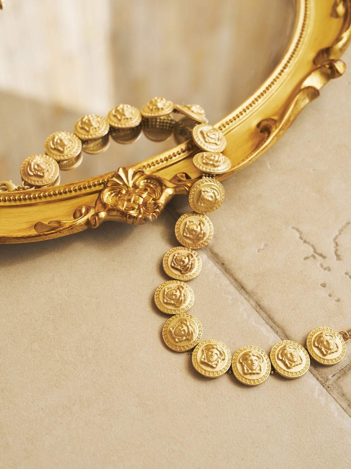 The Social Queen Baroque Medallion Necklace is draped over the edge of an ornate framed mirror, resting on a beige tiled surface.