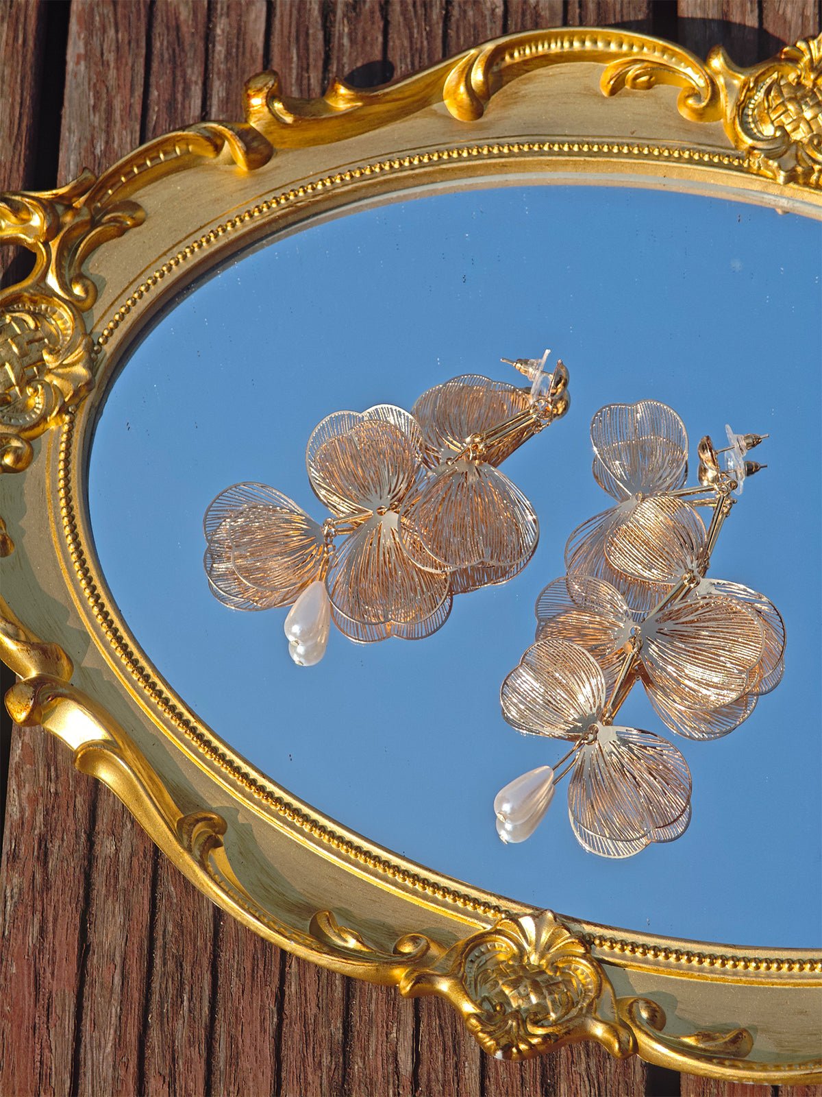 The Baroque Petal Pearl Drop Earrings by Social Queen feature gold-tone petals and water-drop pearls, elegantly displayed on an ornate mirror. Perfect as stylish jewelry for wedding guests.