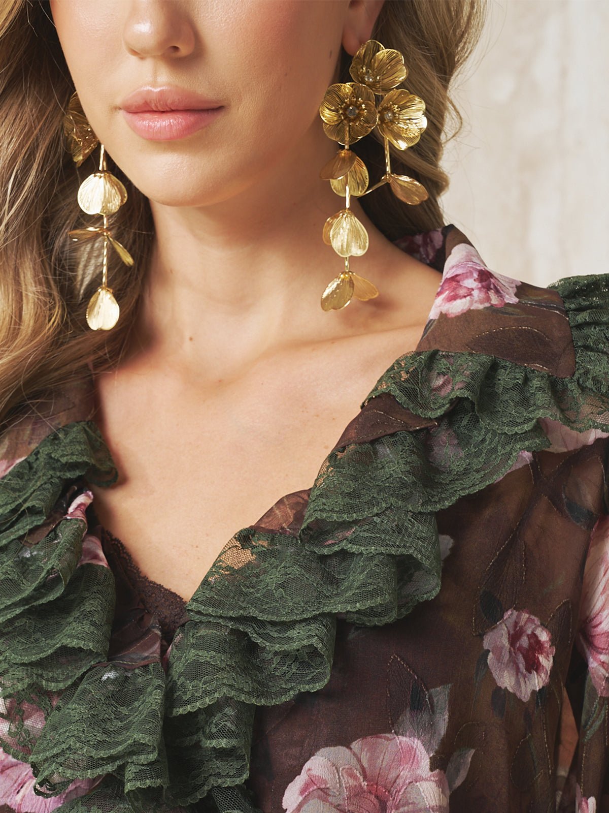 A woman wears Social Queen's Baroque Petal Pearl Drop Earrings with a dark floral top featuring green lace ruffles. Her face is partially visible, and her blonde hair falls in loose waves—a refined pick for wedding guest jewelry.
