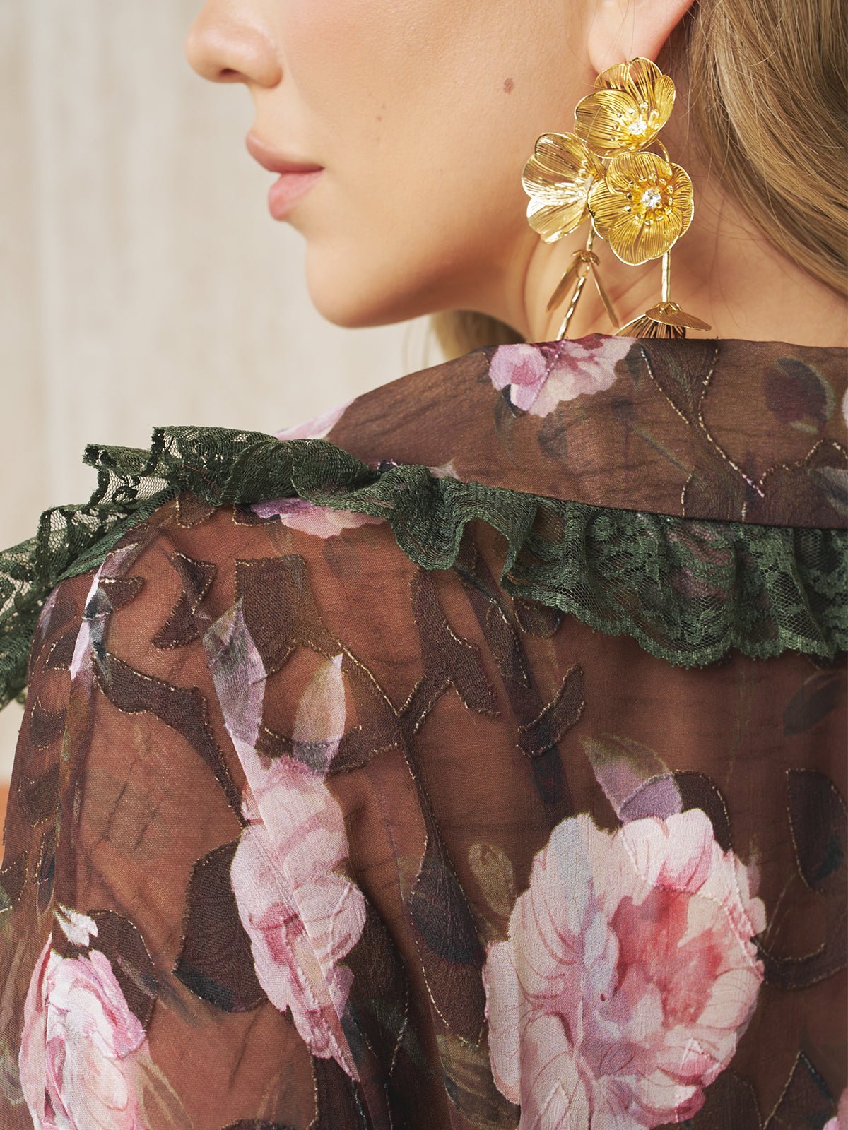 A close-up of a woman wearing a sheer floral blouse with green lace trim and Social Queen Baroque Petal Pearl Drop Earrings, elegant jewelry perfect for a wedding guest.