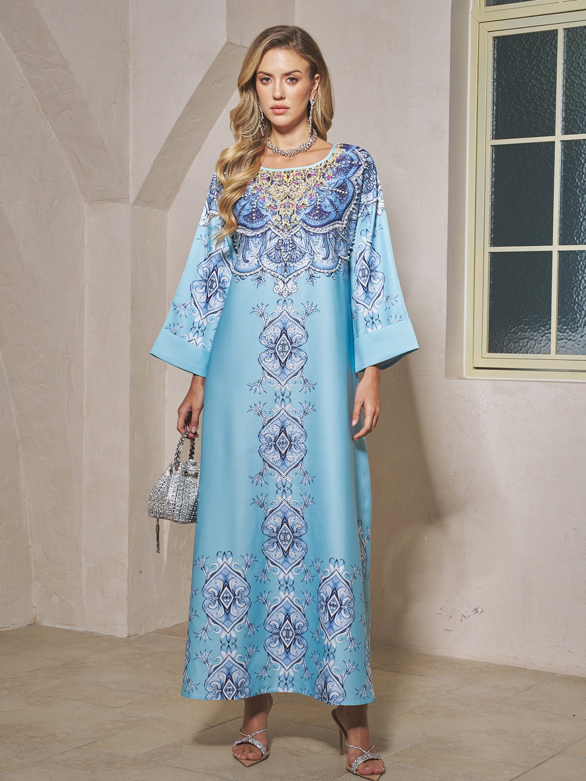 A woman stands indoors wearing the Social Queen Beaded Baroque A-Line Dress in light blue, featuring wide sleeves and a baroque print. She holds a small handbag and pairs the look with silver sandals and jewelry.