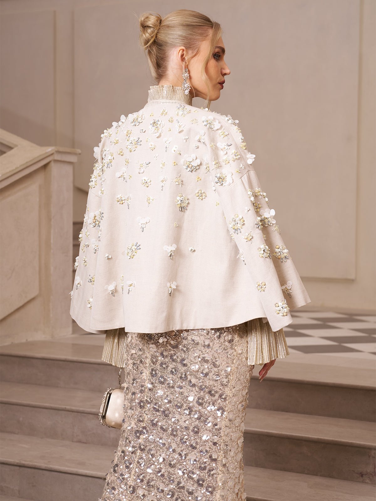 A wedding guest wears the Social Queen Beaded Floral Cape Jacket with 3D floral details and stands on stairs, holding a small purse, her hair in a bun and facing sideways.