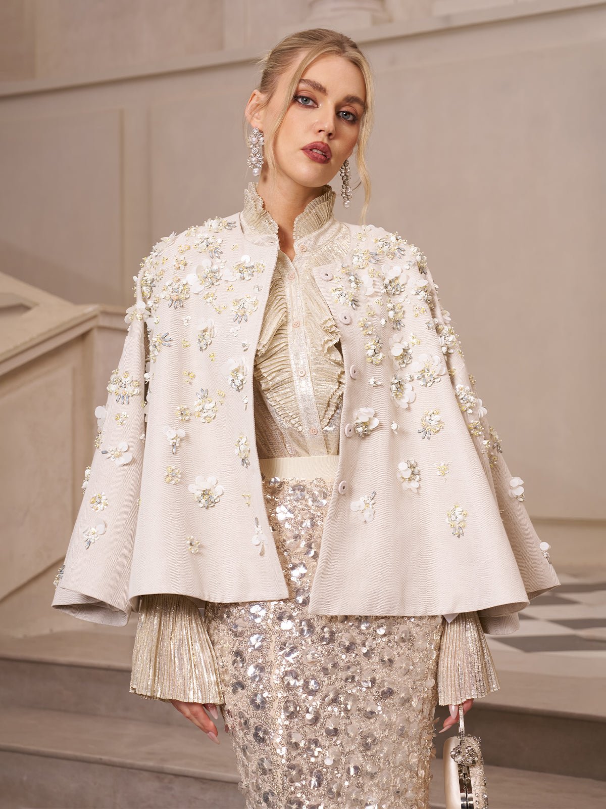 A woman stands indoors by a staircase wearing the Social Queen Beaded Floral Cape Jacket over a metallic sequin dress—a standout wedding guest look.