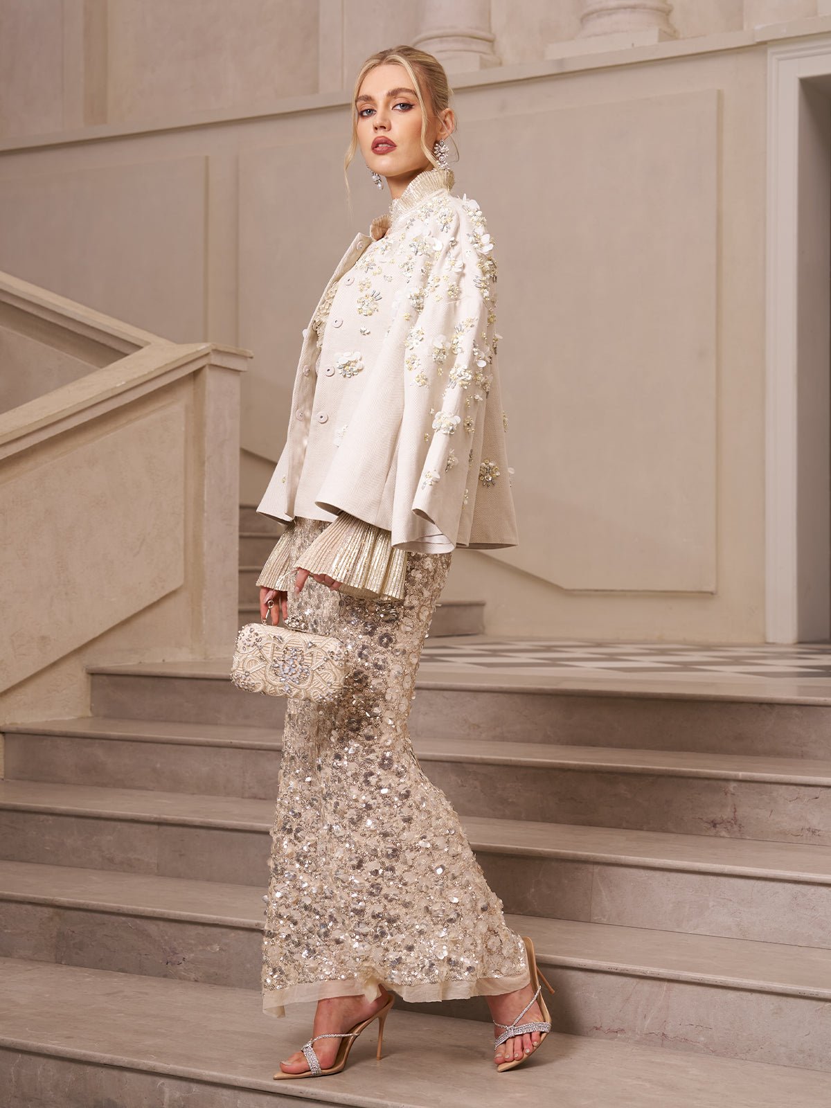 A woman stands on marble stairs wearing Social Queen’s Beaded Floral Cape Jacket, paired with heels and a matching clutch—a chic wedding guest look in an elegant, neutral interior.
