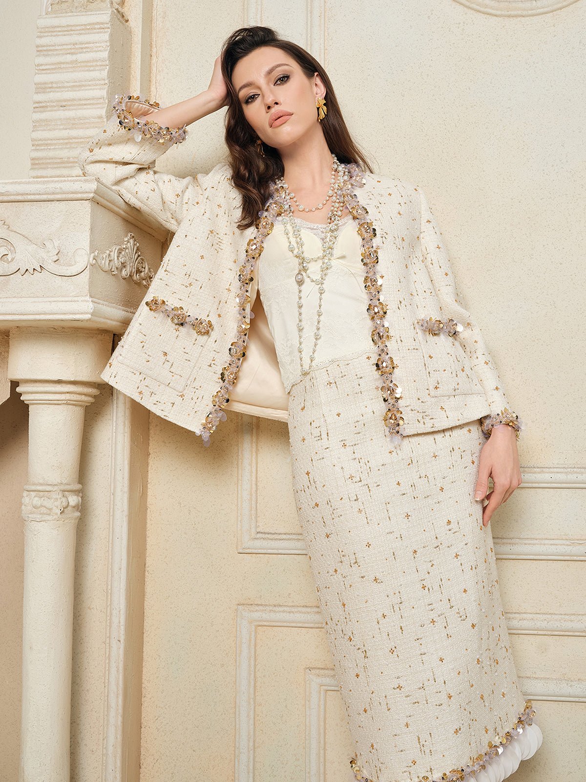 A woman stands indoors in the Social Queen Beaded Floral Embroidered Tweed Jacket, styled with a white blouse and long layered necklaces, leaning against a fireplace mantel.