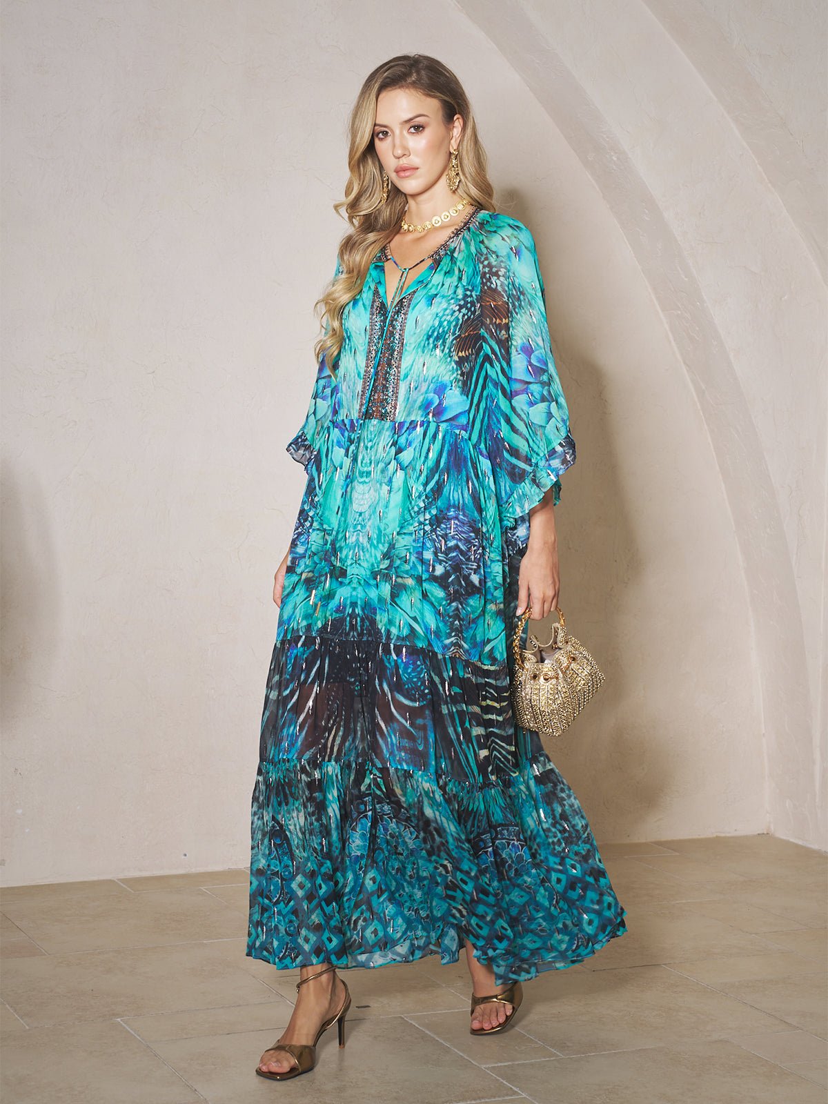 A woman in a Social Queen Beaded Floral Print Ruffled Dress with blue and turquoise beads holds a small handbag indoors, standing on beige tiled flooring against a light-colored wall.