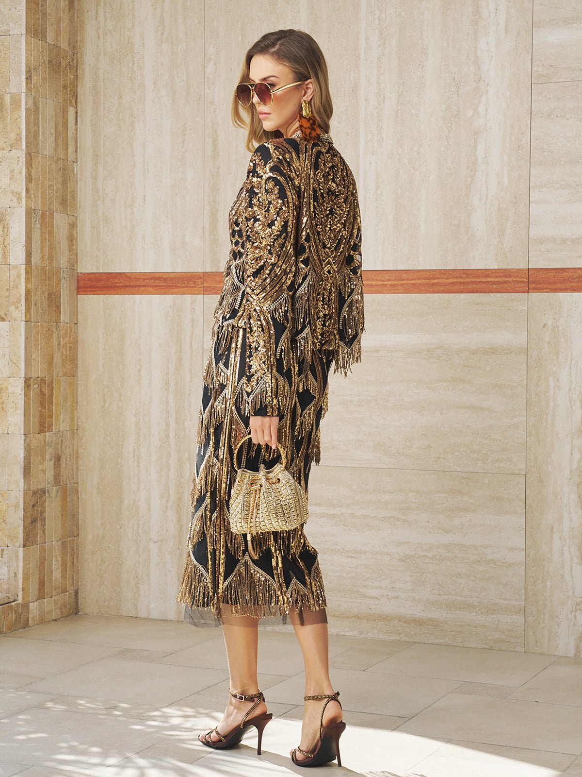 A woman stands indoors by a tiled wall wearing the Social Queen Beaded Fringe Midi Skirt, a black dress with gold details and geometric fringe, paired with sunglasses, high-heeled sandals, and a small gold handbag.