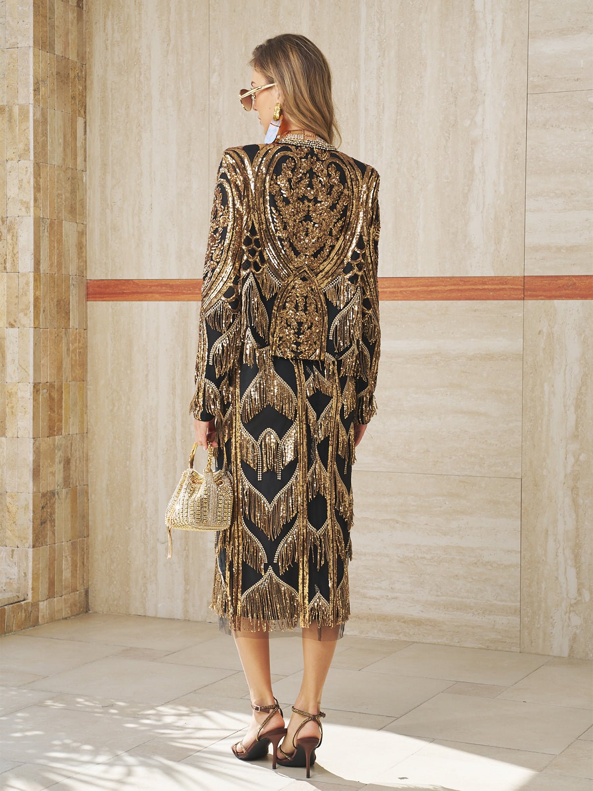 A woman stands facing away in the Social Queen Beaded Fringe Midi Skirt, featuring intricate gold beadwork and a geometric fringe, paired with a small handbag and heels in a light-colored, tiled space.