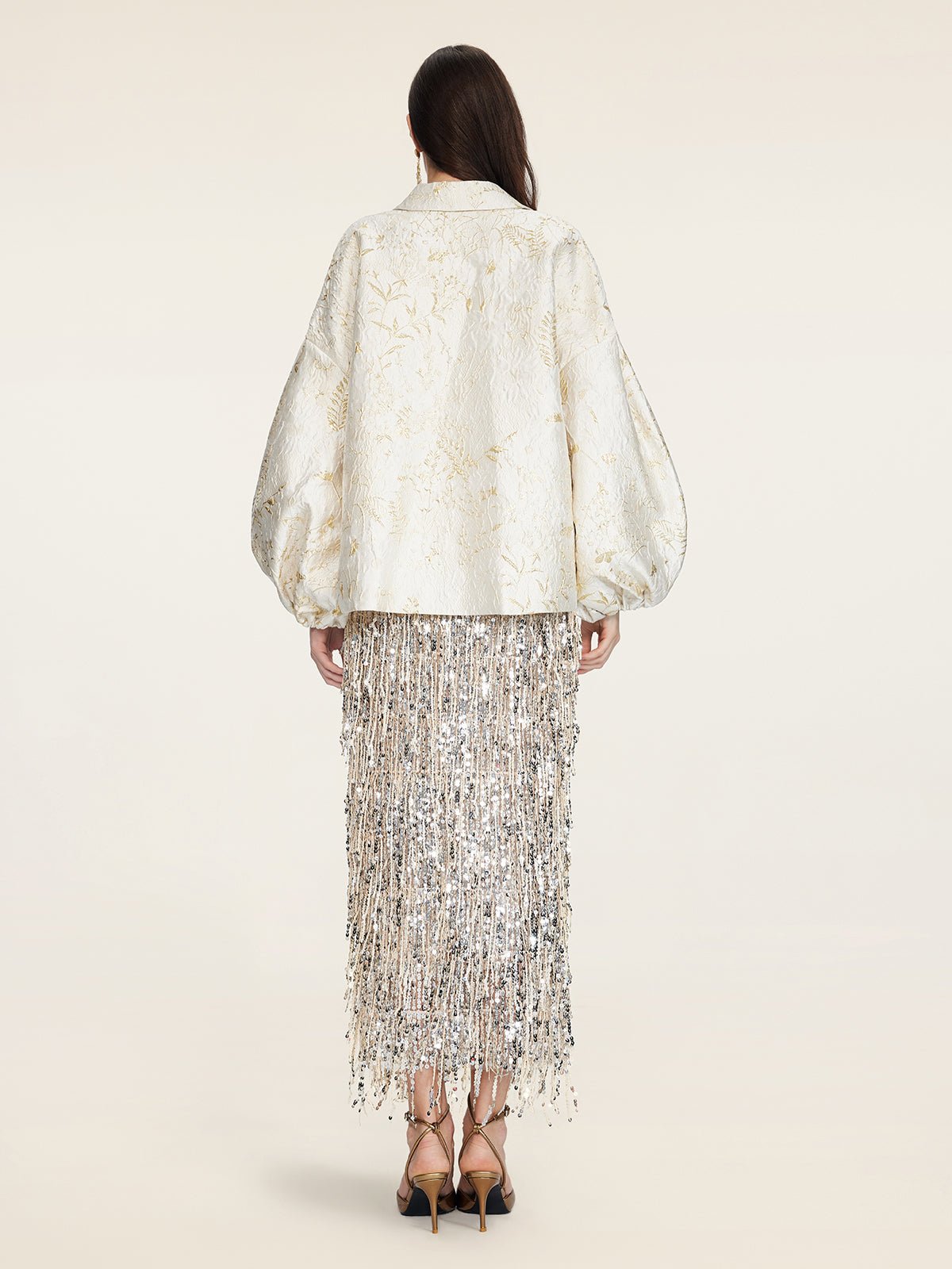 A woman faces away, wearing the Social Queen Beaded Jacquard Lantern Sleeve Jacket in cream, paired with a silver fringed hand-beaded skirt and black high-heeled sandals.