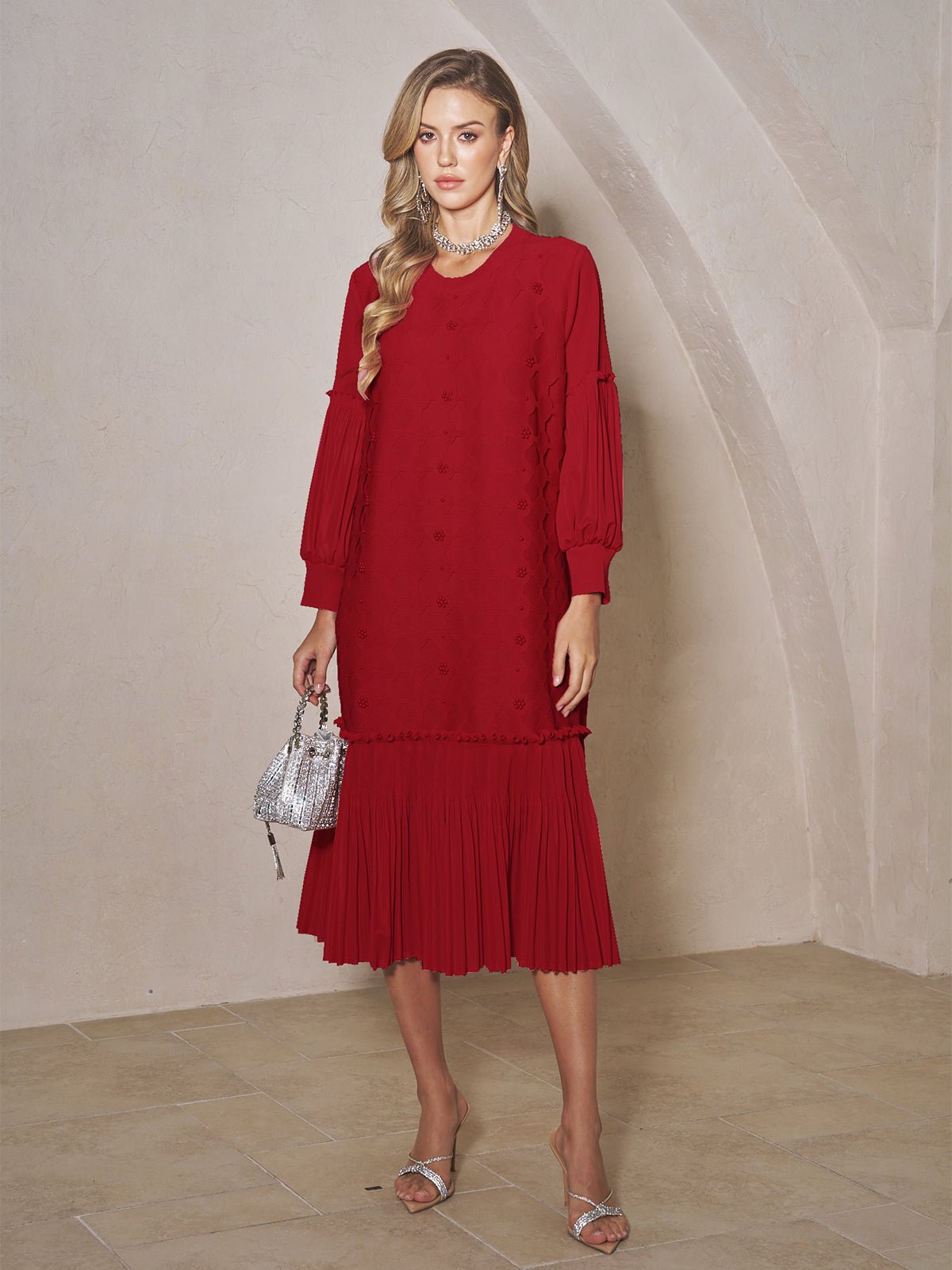 A woman stands indoors against a beige wall, wearing Social Queen's Beaded Jacquard Pleated Midi Dress in red with silver sandals, a silver necklace, and a small silver handbag.