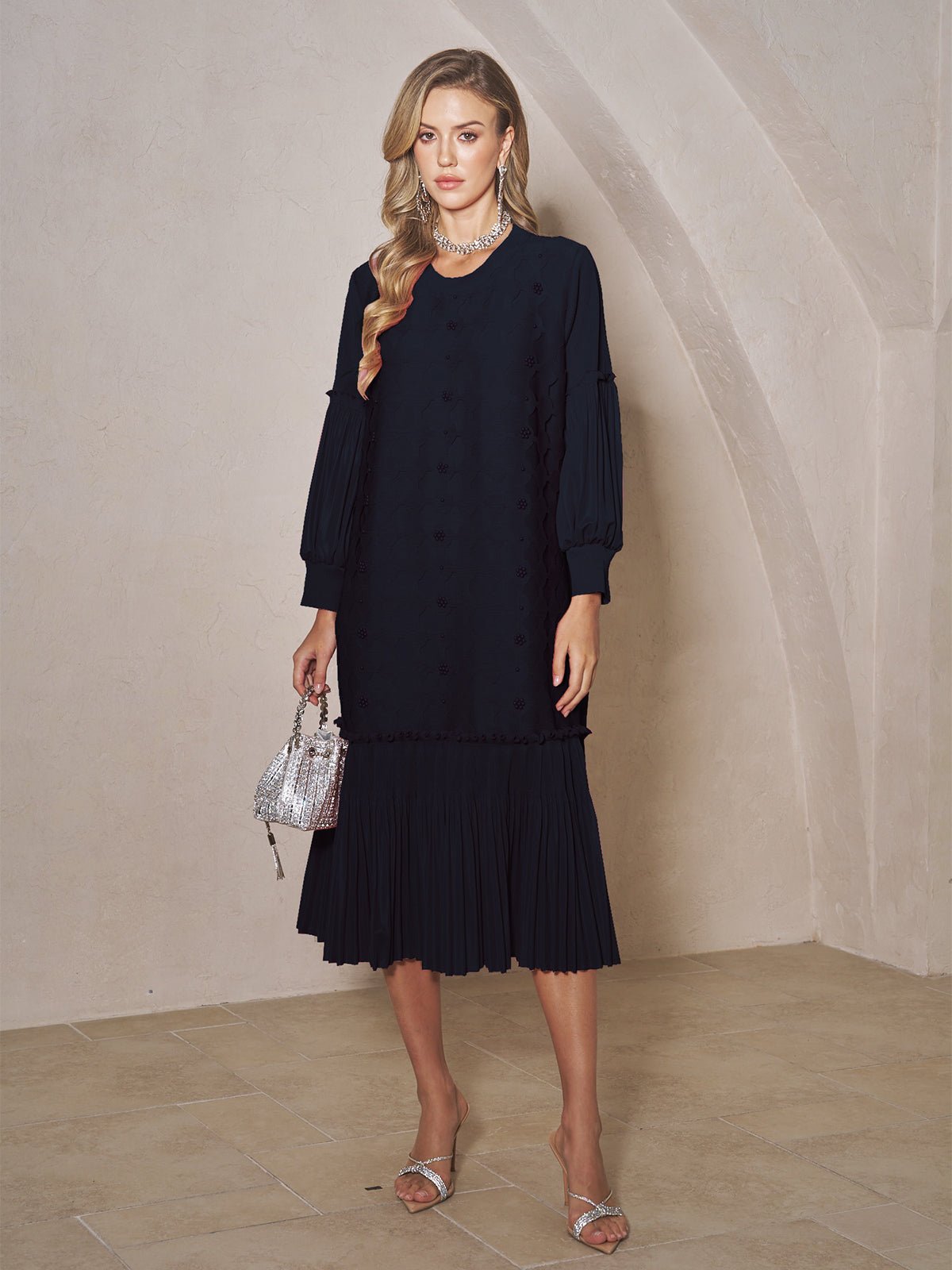 A woman stands indoors wearing the Social Queen Beaded Jacquard Pleated Midi Dress, styled with a silver necklace, silver heels, and a small silver handbag.