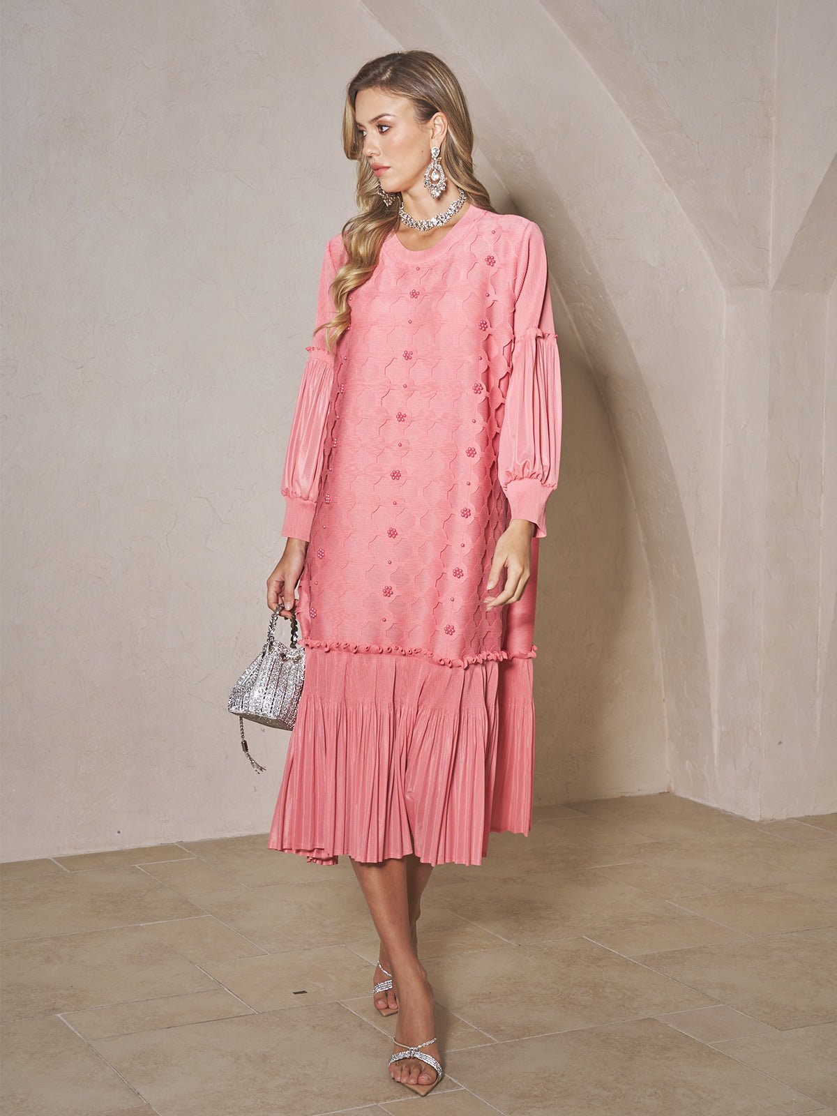A woman in a Social Queen Beaded Jacquard Pleated Midi Dress stands indoors, holding a small silver handbag and wearing silver sandals with statement jewelry.