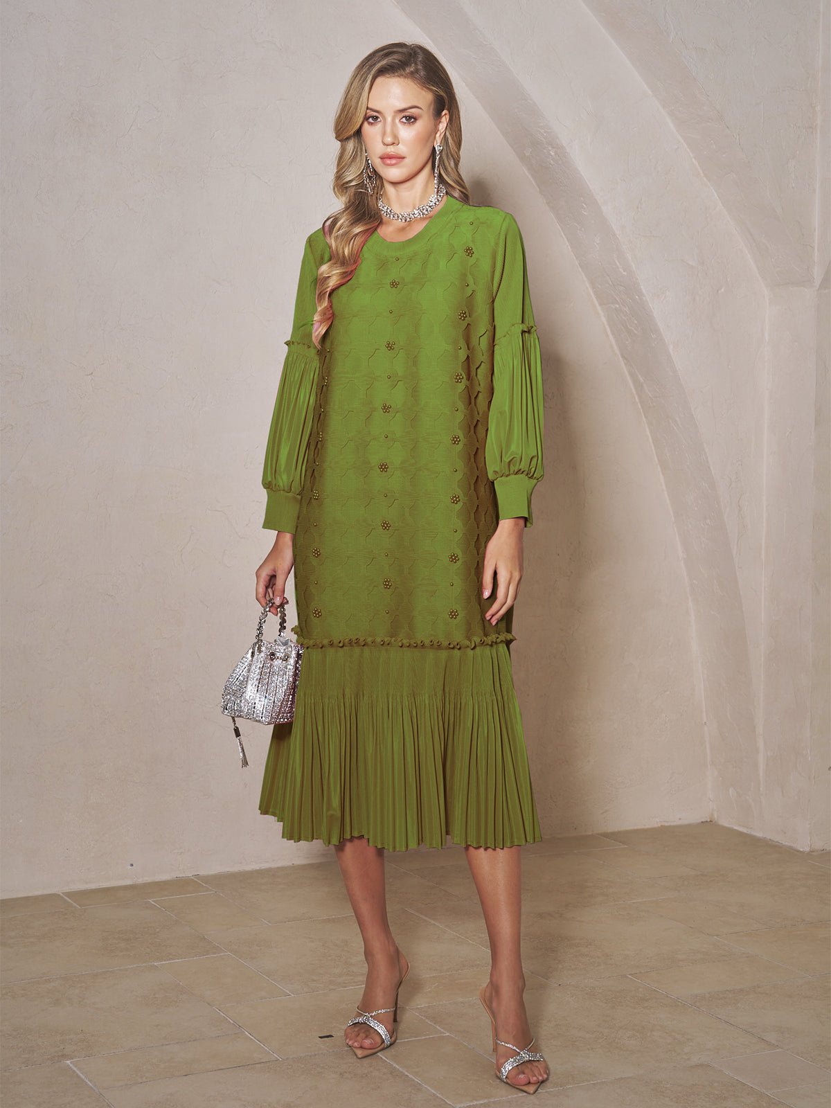 A woman stands indoors in the Social Queen Beaded Jacquard Pleated Midi Dress with silver sandals and a small silver handbag. The arched, light-colored background enhances the elegance of her pleated skirt.
