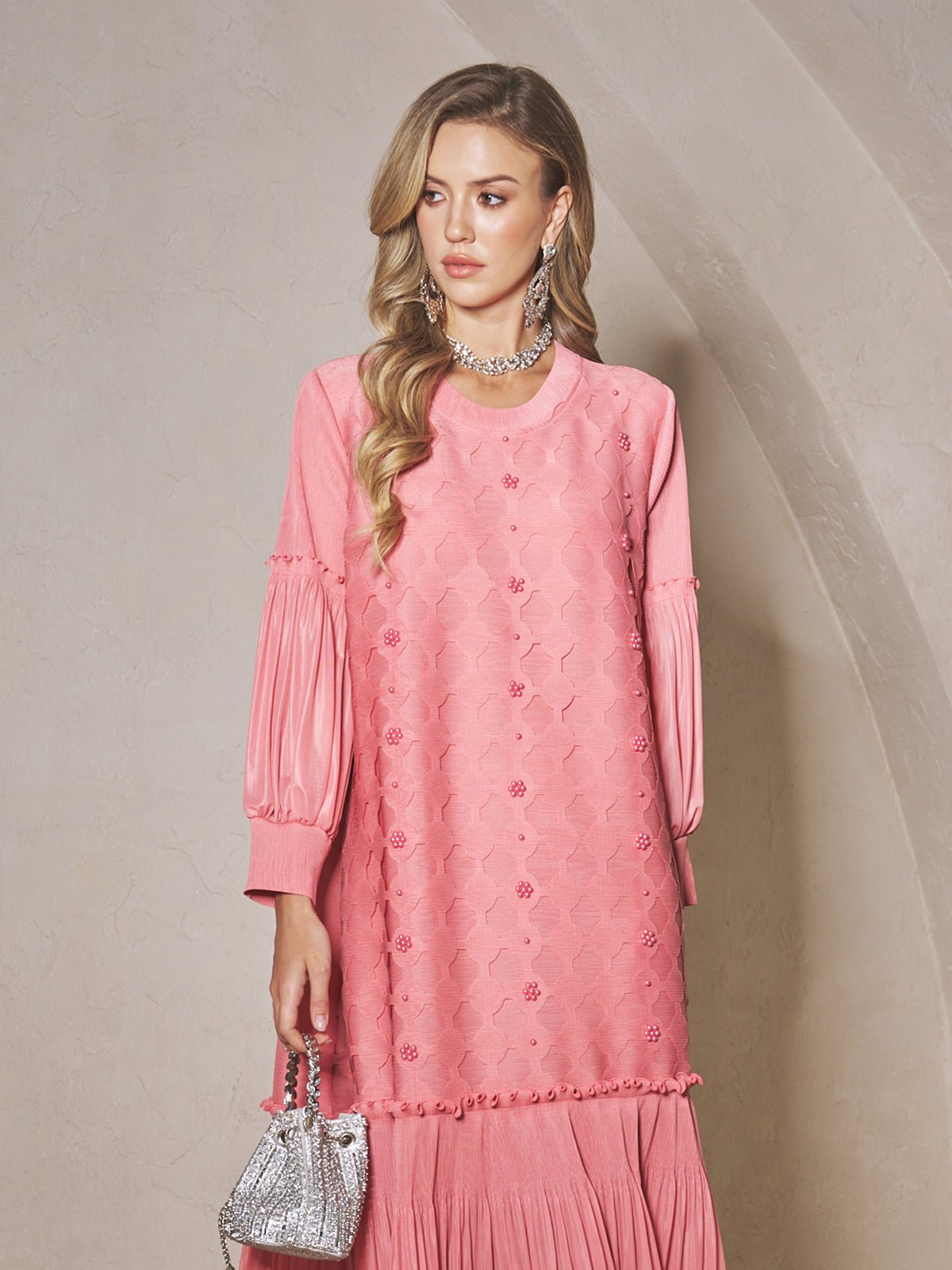 A woman wears the Social Queen Beaded Jacquard Pleated Midi Dress in pink, paired with a small silver handbag and a statement necklace, standing against a light-colored, curved wall.