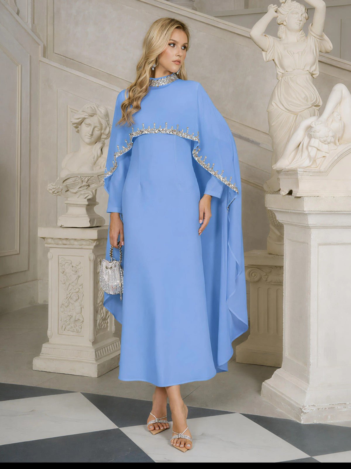 A woman stands indoors near classical statues and a staircase, wearing the Social Queen Beaded Mock-Neck Cape Dress in light blue and holding a small silver handbag.