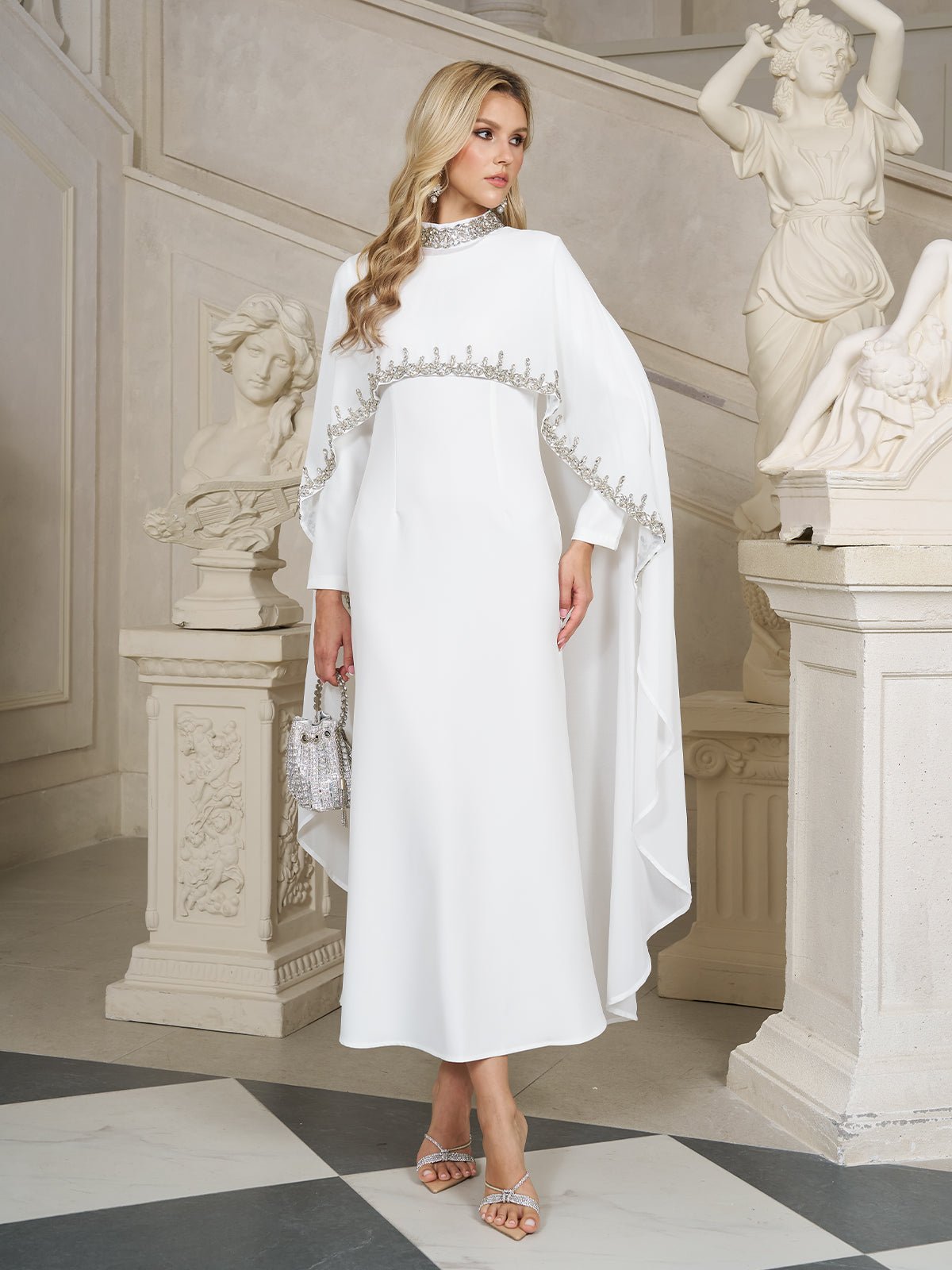 A woman in Social Queen's Beaded Mock-Neck Cape Dress stands indoors by classical statues and a staircase, holding a small silver handbag.