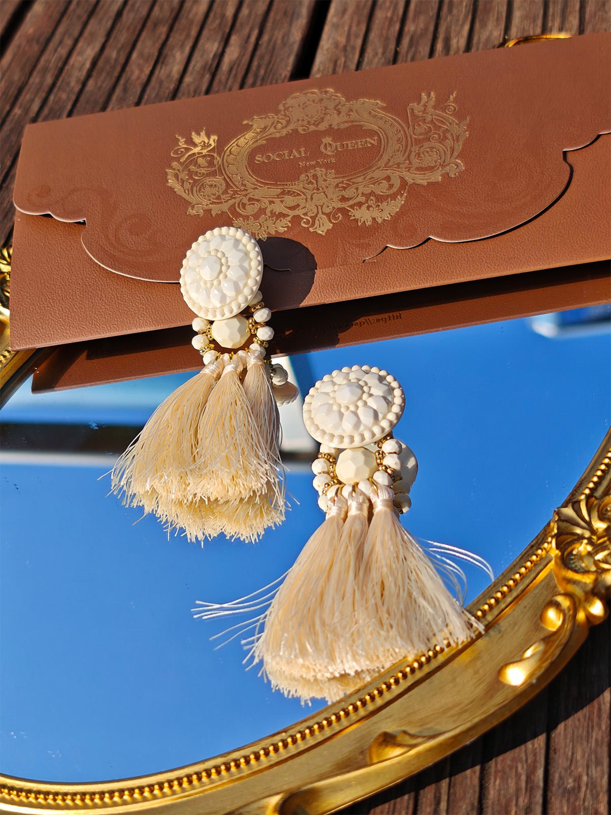 Beaded Tassel Earrings with Silk Fringe by Social Queen in cream rest on a gold-framed mirror, with a brown ornate envelope behind—an elegant choice for wedding guest jewelry.