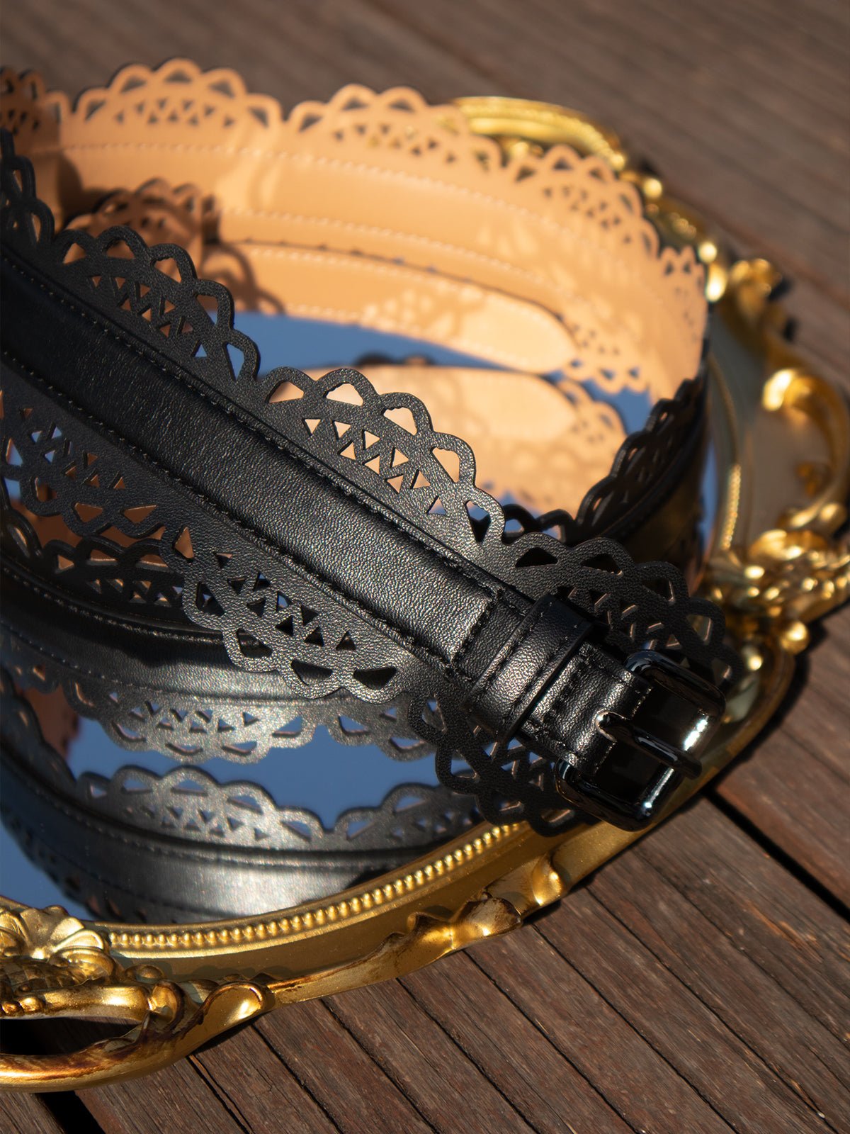 Social Queen's Bohemian Laser-Cut Leather Belts in black and beige, featuring scalloped edges, are showcased coiled on a decorative gold tray atop a wooden surface.